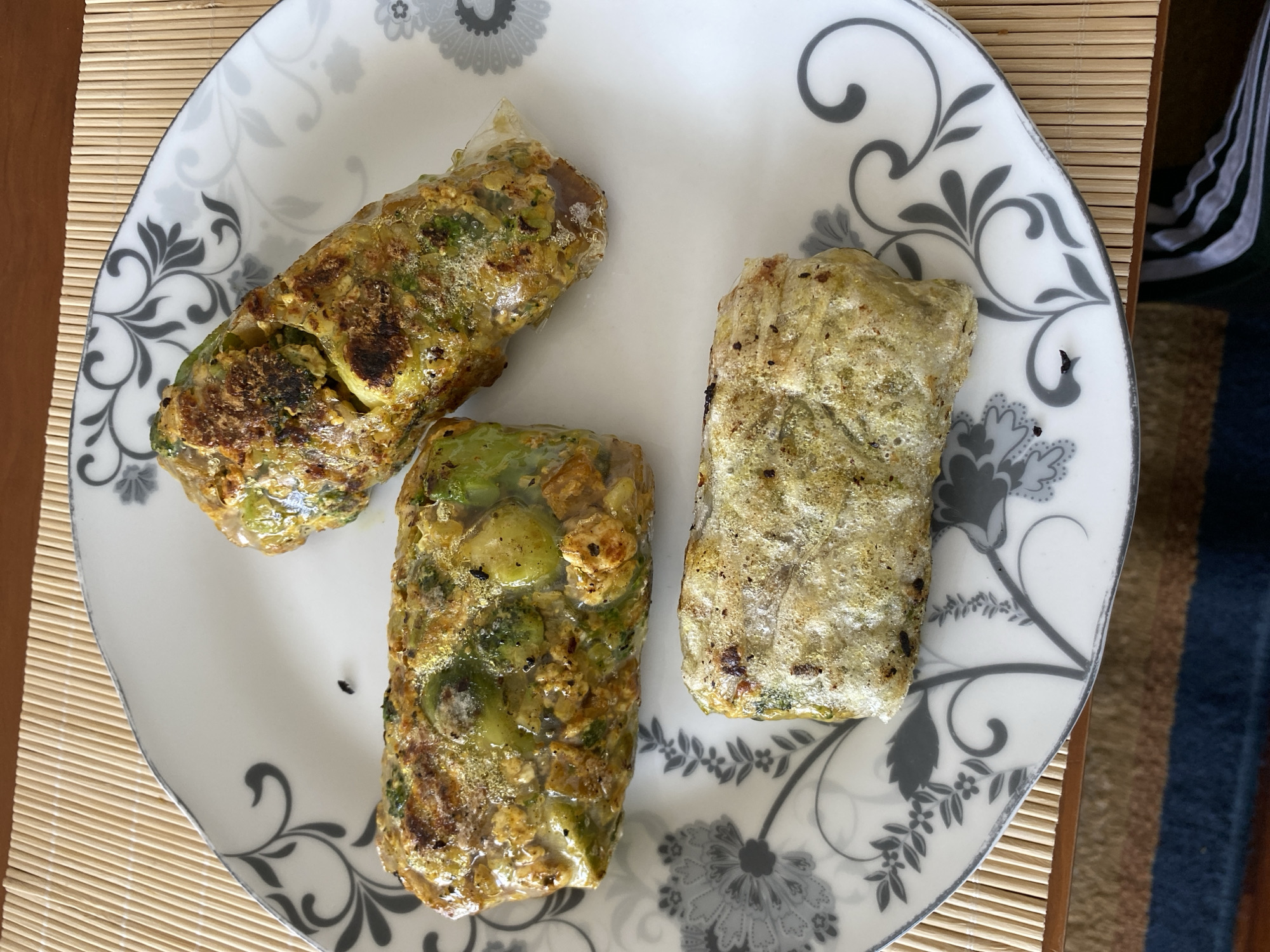This image captures a close-up, overhead view of three rectangular food items served on a white plate with a decorative grey floral pattern. The food items resemble spring rolls or summer rolls, each encased in a translucent, slightly golden-brown wrapper, suggesting they may have been fried or pan-seared. Through the wrapper, a visible filling consists of distinctly green, chunkier vegetables, possibly broccoli or Brussels sprouts, mixed with lighter, granular ingredients that could be scrambled egg or crumbled protein.
The plate rests on a light-colored bamboo placemat, composed of thin, horizontal strips. In the background, beyond the plate and placemat, parts of a dark blue and brown striped rug or mat are visible, along with a wooden surface, indicating an indoor setting, likely a dining area. The lighting appears natural, suggesting the photograph was taken during the daytime. There are no people visible, no specific activities beyond the presentation of food, and no visible text in the image.