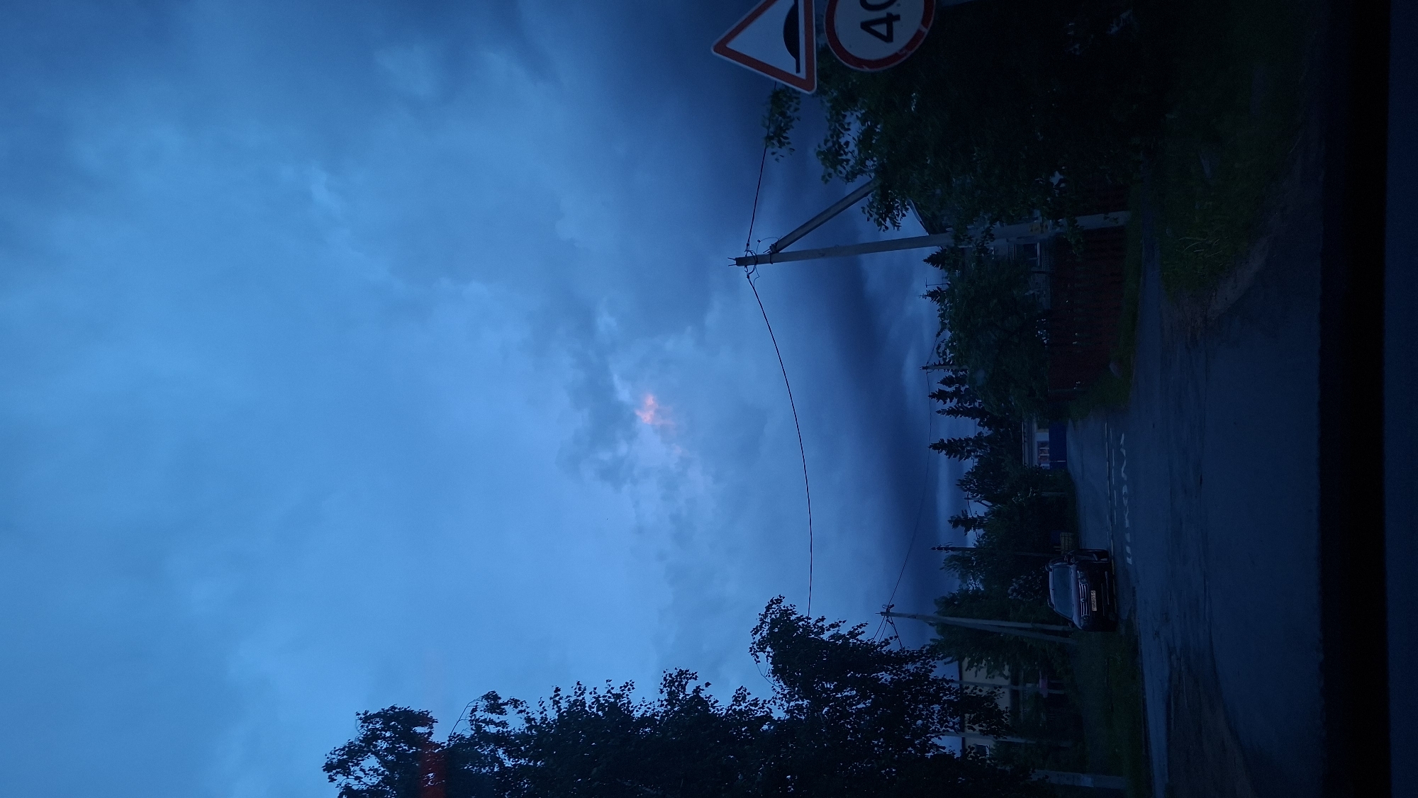 The image depicts a street scene in Забалоцкі сельскі Савет, Belarus, taken at dusk or night. The sky is predominantly blue with dark, heavy clouds, suggesting an impending storm. A small patch of lighter pinkish-orange sky is visible through the clouds. In the upper right corner, a road sign showing "40" is partially visible. A utility pole with wires runs diagonally across the frame. Trees and vegetation line the street on the right side. A car is parked on the right side of the road. The road itself appears to be wet and dimly lit. The overall impression is one of a quiet, rural or suburban street on a cloudy evening.