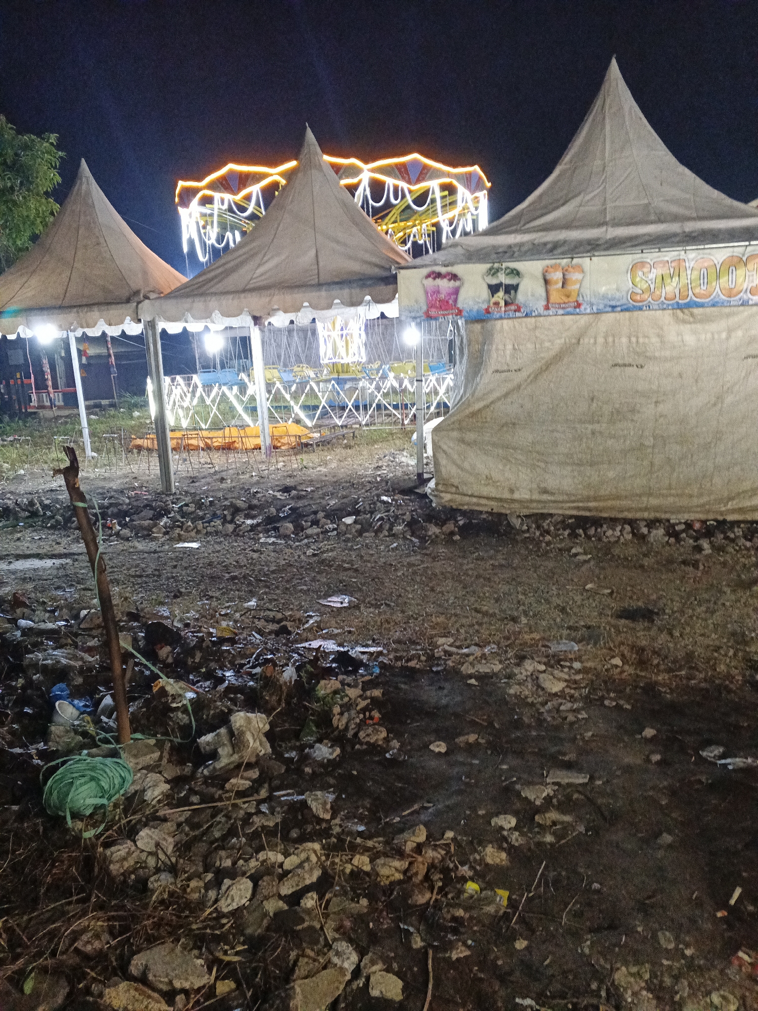 The image depicts a nighttime scene at what appears to be an outdoor event or fairground. In the foreground, the ground is covered in debris, including rocks, dirt, and various forms of trash. A green coil of rope lies on the ground near a wooden post.
Behind the debris, several large, light-colored tents with pointed roofs are set up. One tent has a banner advertising "SMOOTH" with images of colorful drinks. The tents are illuminated from within by bright lights.
In the background, a brightly lit amusement ride, possibly a carousel or Ferris wheel, is visible against the dark night sky. The ride is adorned with numerous orange and white lights, creating a festive atmosphere. The overall environment appears somewhat unkempt, with the debris in the foreground contrasting with the illuminated structures and ride in the background. The location is identified as Sukabumi, Indonesia.