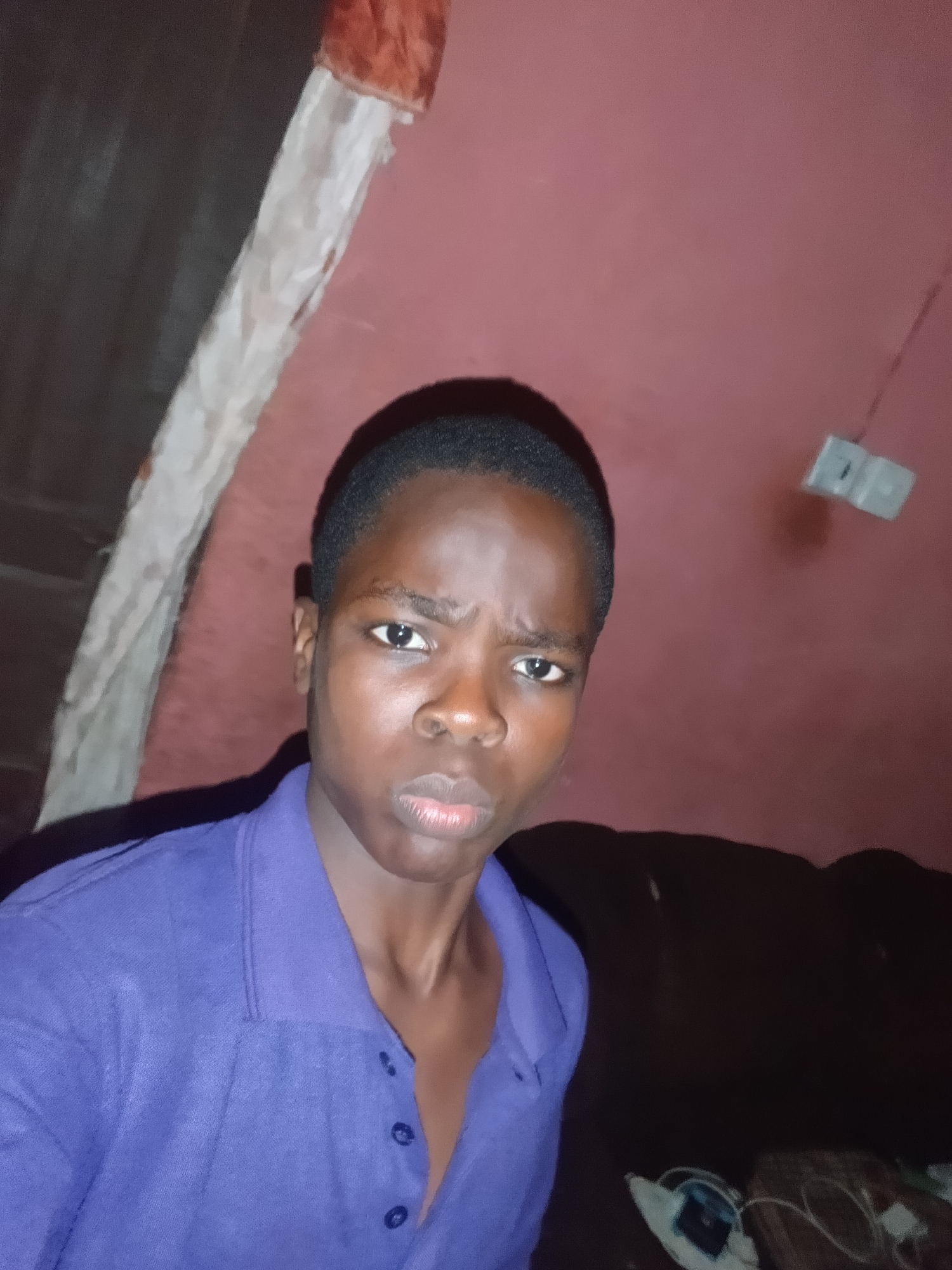 A young Black male, positioned centrally from the chest up, looks directly into the camera with an intense, slightly furrowed expression and subtly pursed lips. He has short, dark hair and is wearing a dark blue or purple polo shirt with three visible buttons on the placket. The background features a reddish-pink wall that transitions into dark, shadowed areas. A textured, light-colored vertical element, possibly a pipe or structural support, runs diagonally from the upper left. In the upper right, a small, dark rectangular object, possibly an electrical outlet, is mounted on the reddish-pink wall. The lower right background is dark, suggesting furniture, with indistinct objects and cables visible at the bottom right. The scene is illuminated indoors. This image depicts a location in Onitsha, Nigeria.