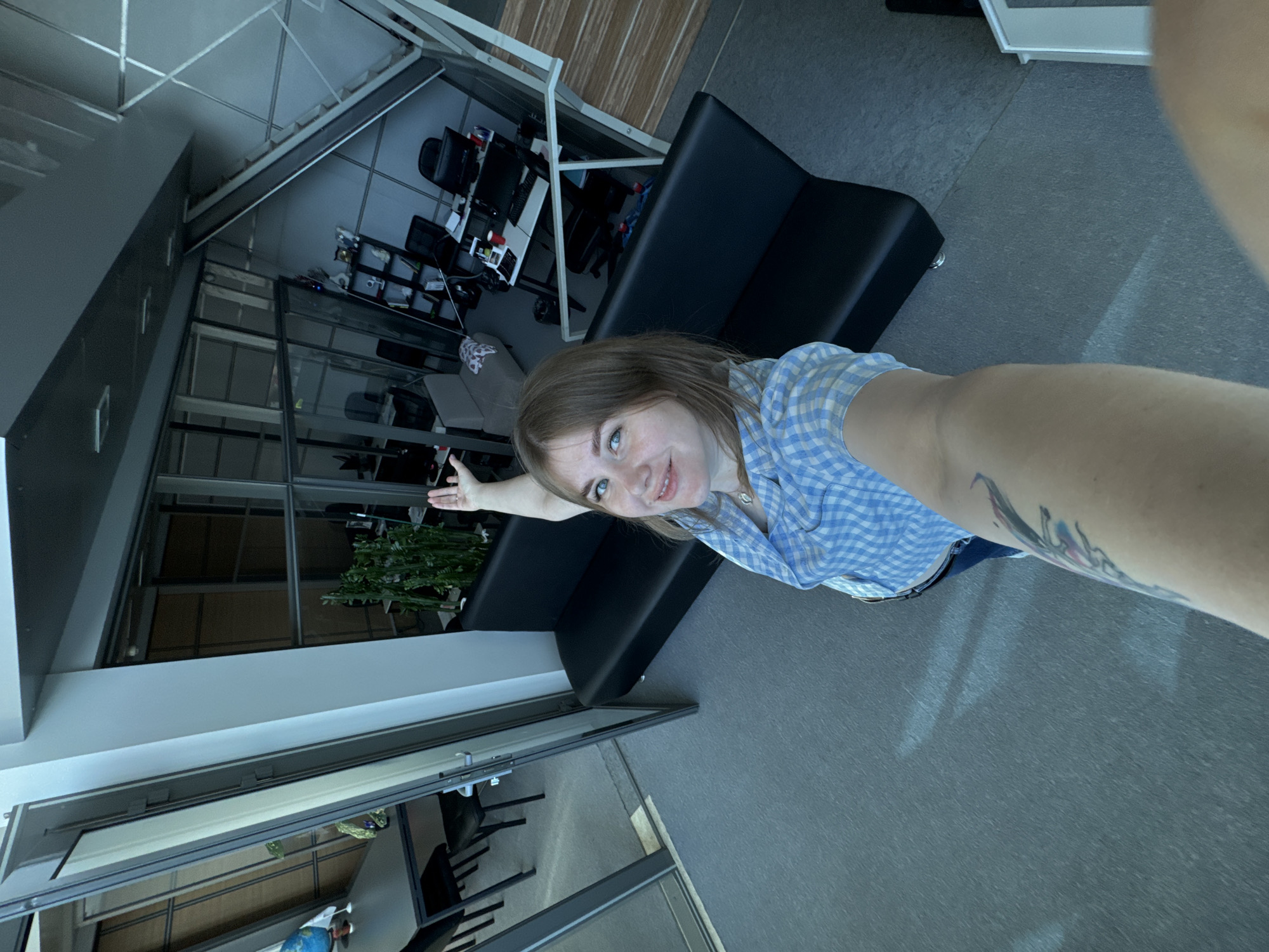 The image shows a woman with shoulder-length brown hair, wearing a blue and white checkered shirt, taking a selfie in what appears to be an office space. She is smiling and gesturing to her left with her left arm. A tattoo is visible on her right forearm.
Behind her is a black sofa positioned in front of a glass partition wall. Beyond the glass wall is another office area containing desks, chairs, computers, and filing cabinets. A plant is visible by the glass wall. The floor is a gray, smooth surface. The ceiling has a modern design with intersecting beams.
Based on the camera angle and perspective, the woman is likely standing on a flat surface, holding the camera high and to her right.
It is not possible to verify that this photo was taken in Perm, Russia.