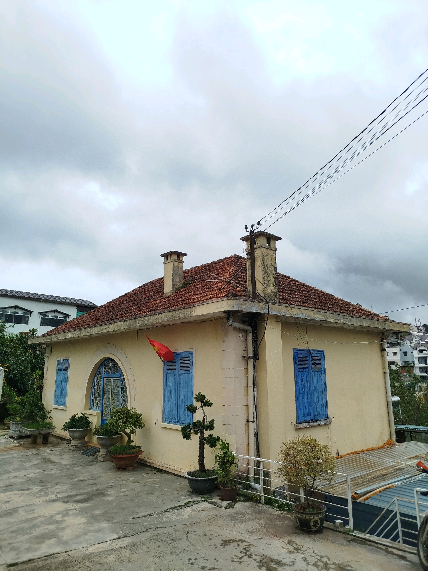 The image depicts a light yellow, single-story house with a red-tiled roof and blue shutters on its windows, located in Da Lat, Vietnam. A Vietnamese flag is visible, attached to the wall near a chimney. The house has a distinctly rounded archway above its front door. Potted bonsai trees and other plants are strategically placed around the exterior, adding greenery to the scene. The house appears to be situated on a slightly elevated lot, with other buildings visible in the background, suggesting an urban or suburban setting. The sky is overcast with heavy clouds, indicating a possibly cloudy or rainy day. The ground in front of the house is paved with concrete, showing some cracks and weathering. Electrical wires are strung across the upper portion of the image, typical of Vietnamese streetscapes.