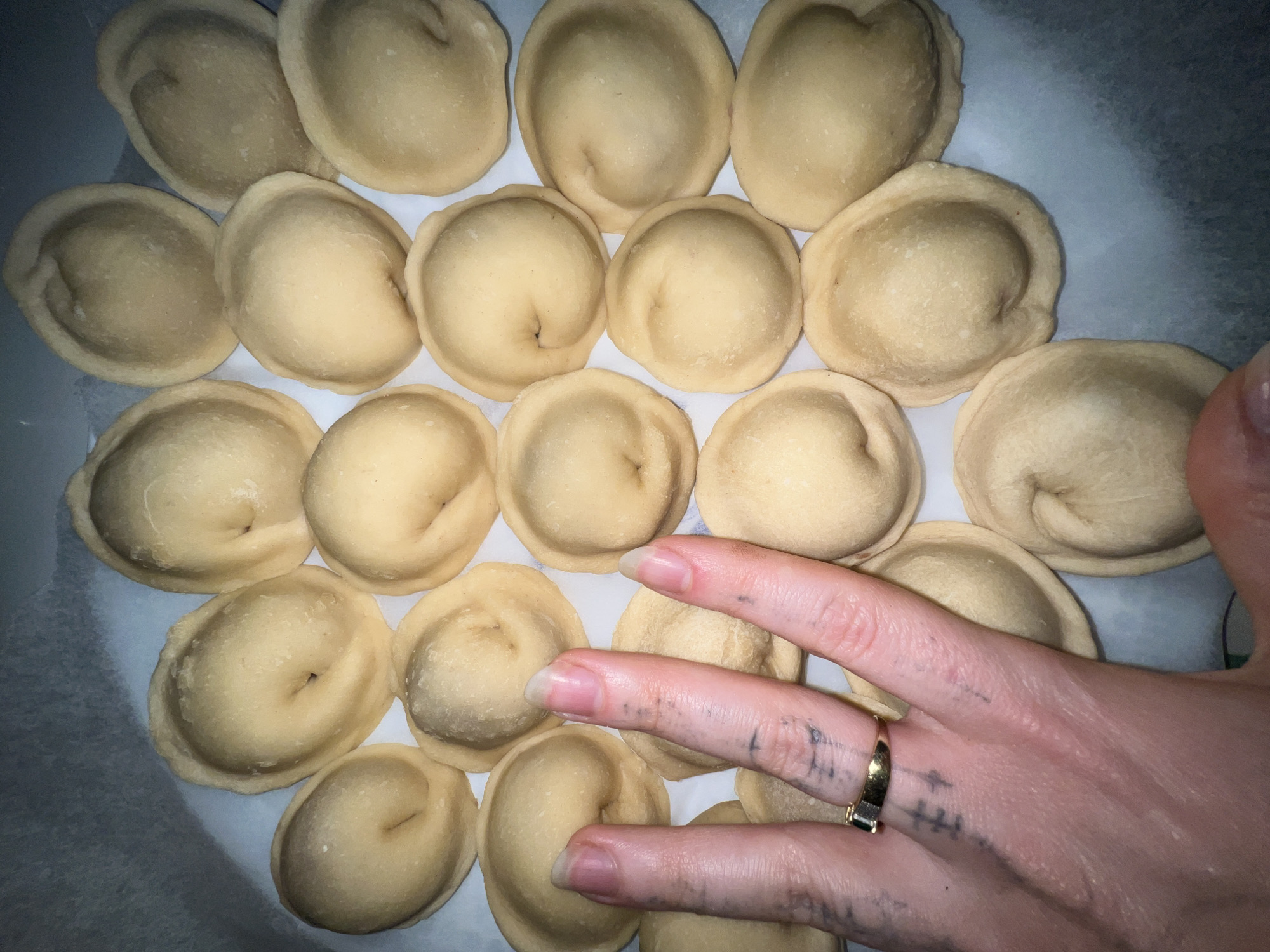 A close-up shot depicts approximately twenty raw, circular dumplings arranged in a pattern on a white surface or tray. A hand with a gold ring on the ring finger is also in the frame, overlapping some of the dumplings. The hand has light skin, long nails, and what appear to be faded, dark tattoos. The scene suggests a domestic setting, likely a kitchen, in the city of Palilula Urban Municipality, Serbia. The lighting appears to be somewhat artificial.