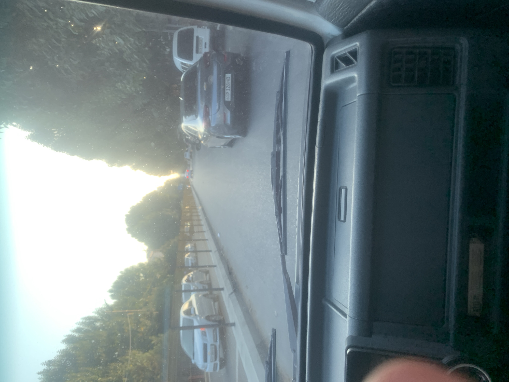 The image is taken from inside a vehicle, looking out through the windshield. The perspective is forward-facing, showing a street scene with trees lining the road. Several cars are visible, parked and in motion. The sun is low in the sky, creating a bright glare and casting long shadows. The dashboard of the vehicle is partially visible in the foreground. The license plate of a gray car in the middle ground reads "60 P 128 HB". The overall environment suggests a suburban or city street during daytime, possibly late afternoon due to the lighting conditions.