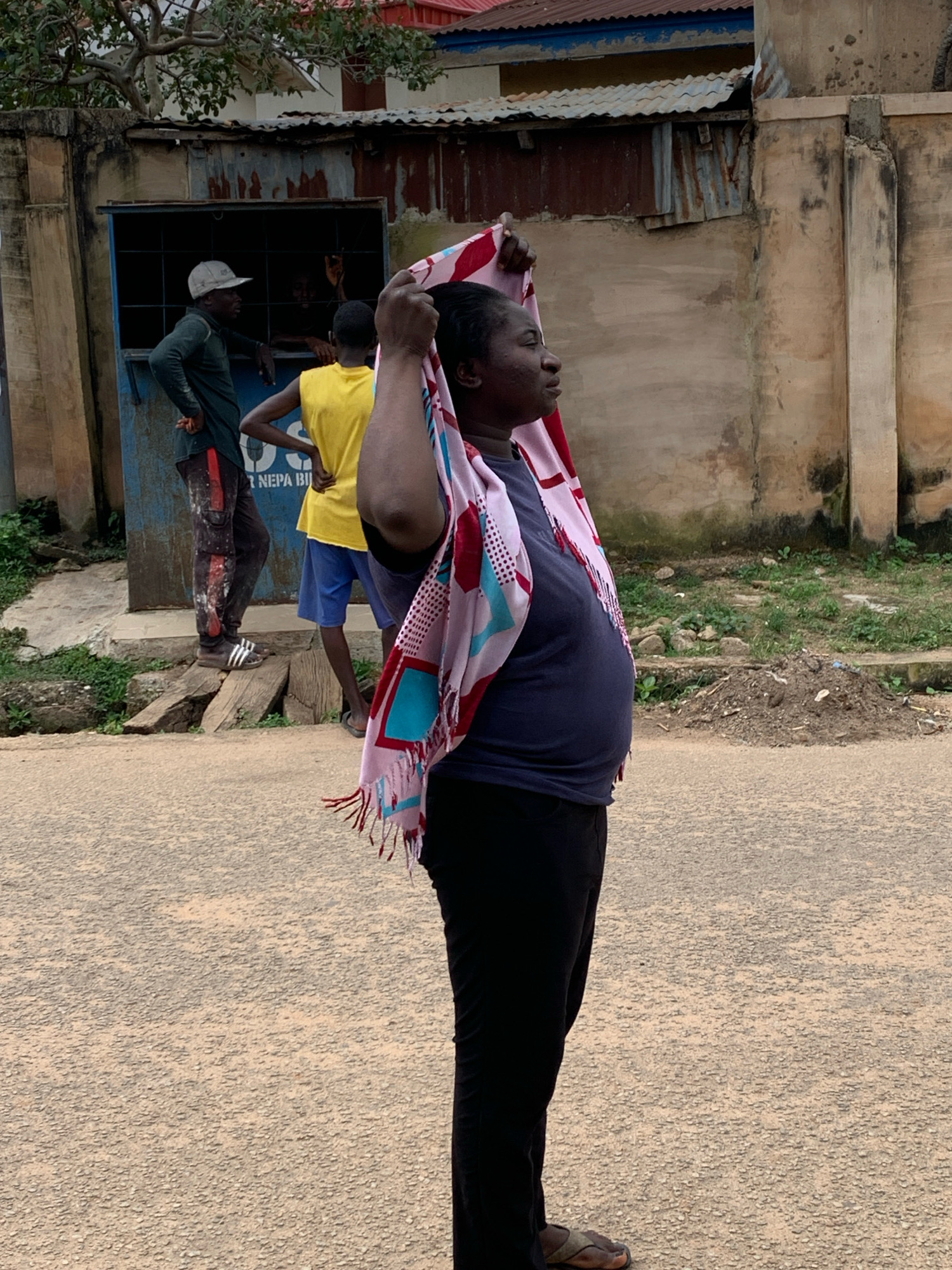 A woman stands in the foreground of the image, her body facing right and her head turned to the side, looking off-camera. She is wearing a dark blue t-shirt and black pants, with a colorful scarf draped over her shoulders and head. The scarf has a geometric pattern in red, blue, and white, with fringed edges. Her right arm is raised, holding the scarf.
In the background, a storefront is visible. Two men and a younger person are standing near the open doorway of the storefront. One man, wearing a dark green long-sleeved shirt and black pants with red stripes, is leaning on the doorway and wearing a light-colored cap. He is looking into the storefront. The younger person, wearing a bright yellow t-shirt and blue shorts, is standing with their hands on their hips, facing away from the camera. Another person is partially visible inside the storefront.
The storefront appears to be made of corrugated metal and painted blue, with bars across the opening. The building behind the storefront has a weathered, pale beige exterior. A partially visible red roof is on top of the building. The ground in front of the storefront and the woman is a dirt or gravel road. There is some sparse vegetation and debris on the ground to the right of the storefront. The overall scene appears to be an outdoor setting in a modest urban environment, likely in Jos, Nigeria, given the architectural style and the presence of the "NEPA BIL" sign on the storefront, which refers to the National Electric Power Authority.