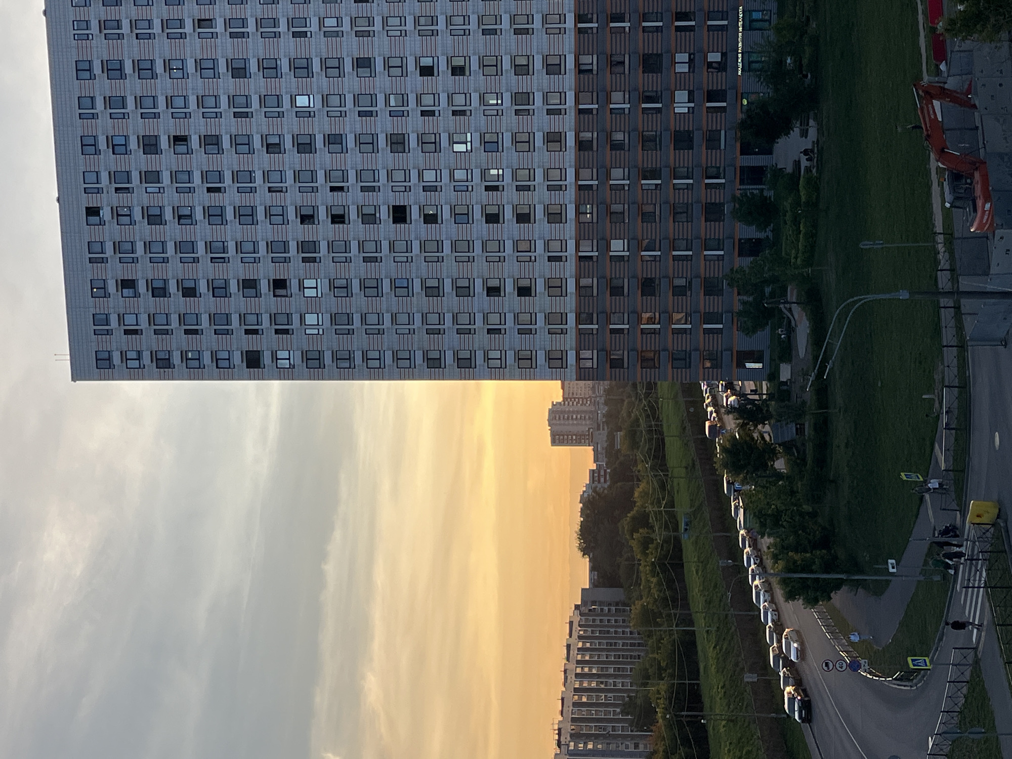 The image depicts a modern residential building in Moscow, Russia, at dusk. The building, which appears to be a high-rise apartment complex, is characterized by its repetitive grid of windows and balconies, with a darker band of color about a third of the way down from the top and another at the bottom. To the left of this building, the sky is a gradient of orange and yellow, indicative of a sunset. Further to the left, other residential buildings and trees are visible, with a road lined with cars curving through the foreground. The road has a yellow pedestrian crossing, and several individuals are walking on the sidewalk. In the lower right corner of the image, a construction site is visible with an excavator. The scene suggests a typical urban evening in a residential area of Moscow.