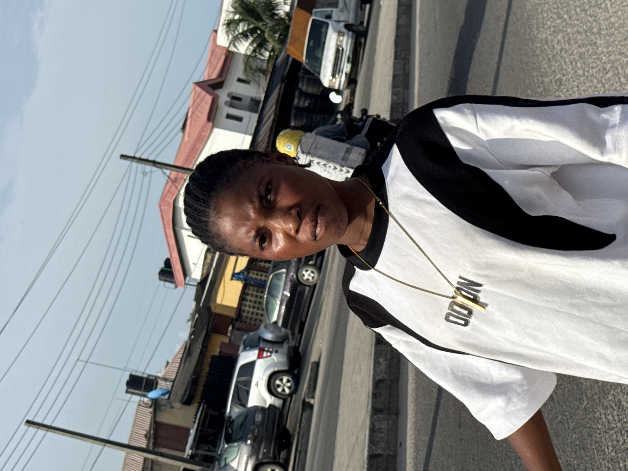 A medium shot depicts a person with short, dark hair wearing a white t-shirt with black accents and a gold necklace. The t-shirt has a word on it, partially obscured. The person is standing outside on what appears to be a city street. The background shows buildings of varying heights and designs, some with red roofing. Cars are parked along the street, with one white van prominent in the background. Power lines stretch across the sky above the buildings. Based on the prompt and image context, the location is in Ado, Nigeria.