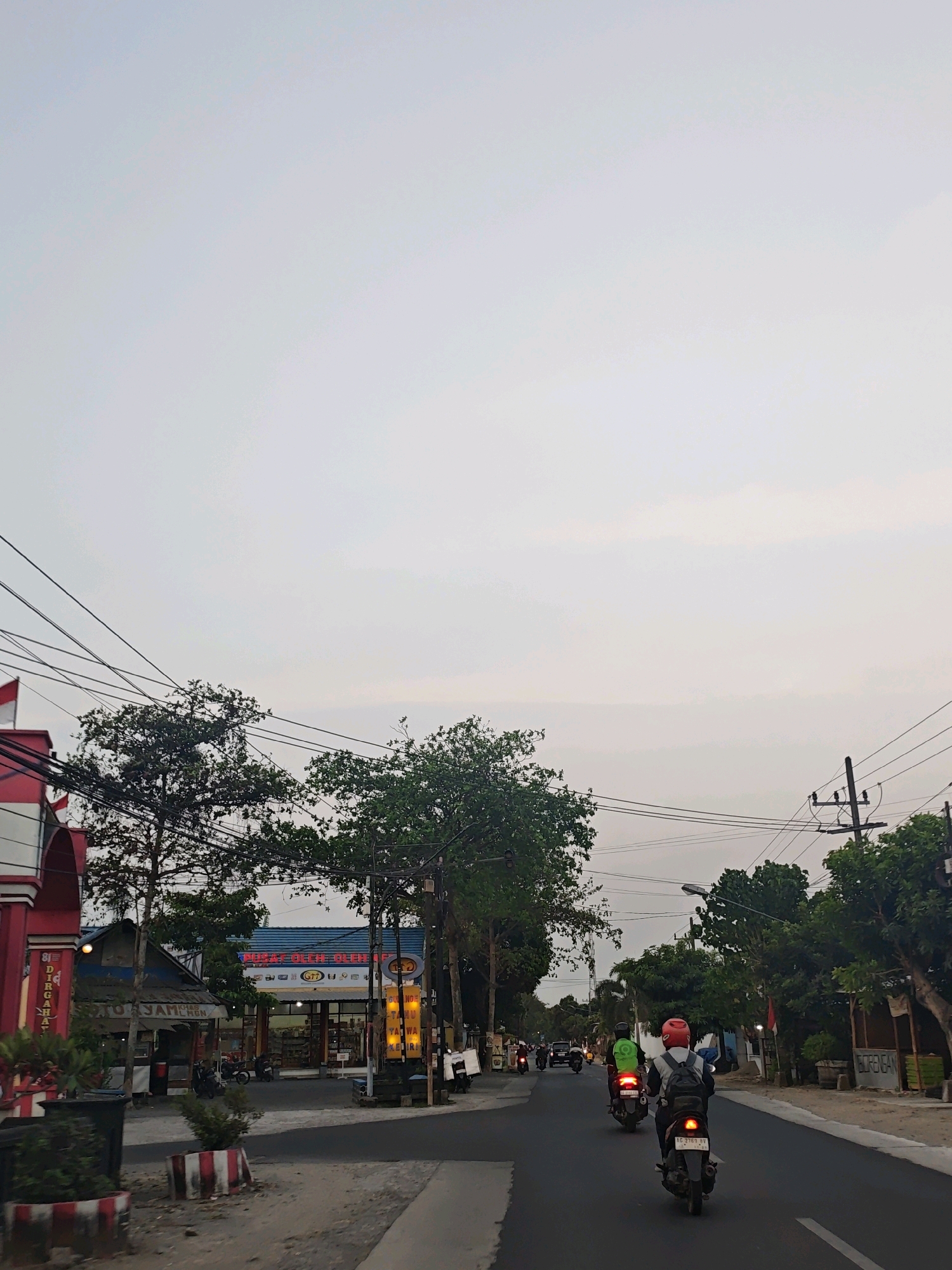 The image captures a street scene in Ngasem, Indonesia, during what appears to be late afternoon or early evening due to the soft, diffused light and overcast sky. A paved road runs through the center of the frame, with several motorcycles visible. In the foreground, a motorcycle with a rider wearing a red helmet and a backpack is moving away from the camera, its taillight illuminated. Further down the road, another motorcycle with a rider in a green jacket is visible, also moving away. A third motorcycle is partially obscured behind it.
On the left side of the road, a building with a red and white facade is present, with an arched entrance. Adjacent to it is a smaller, shaded structure with signage. Across the street, a larger building with a blue awning displays various signs, including one that reads "Pusat Oleh-Oleh H." A prominent yellow sign with text in an Indonesian language is also visible. Several trees with dense green foliage line the street, interspersed with utility poles and a complex network of electrical wires. The environment suggests a commercial or mixed-use area within a town or city, with evidence of local businesses and transportation. The overall atmosphere is calm, with no overt signs of activity beyond the movement of vehicles.