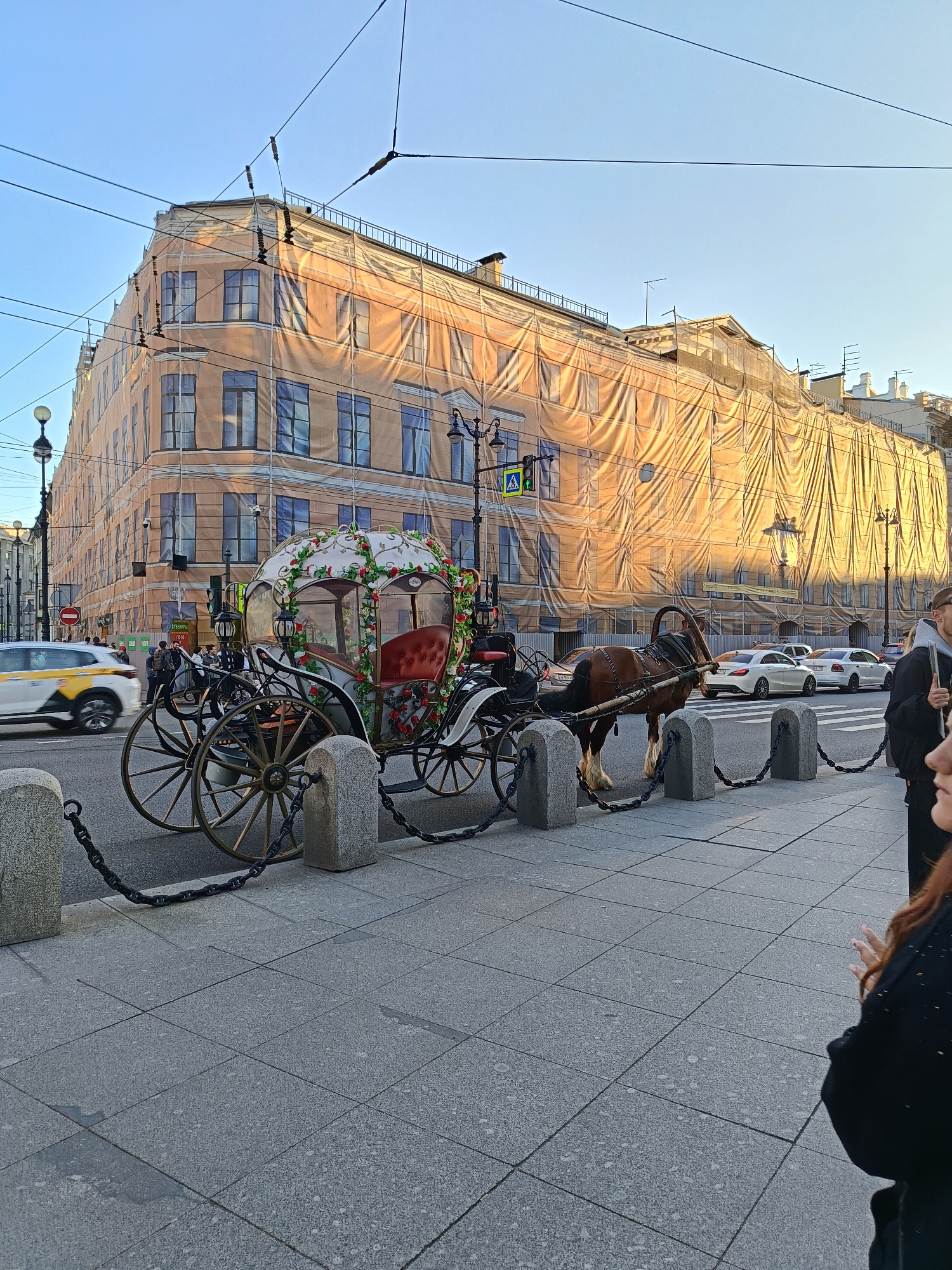 The image shows a horse-drawn carriage parked on a cobblestone sidewalk next to a street in what appears to be a city. The carriage is decorated with white trim and red and green floral accents. A brown horse is hitched to the carriage. The carriage is secured to gray stone bollards by black chains.
In the background, a large building is visible, covered in what appears to be a protective fabric or tarp, possibly for renovation work. There are windows visible behind the covering. A street with multiple vehicles and some pedestrians is located in front of the building.
On the right side of the image, the partial view of a person with red hair is present. The sky is a clear, light blue. Street lights and tram lines can be seen overhead.