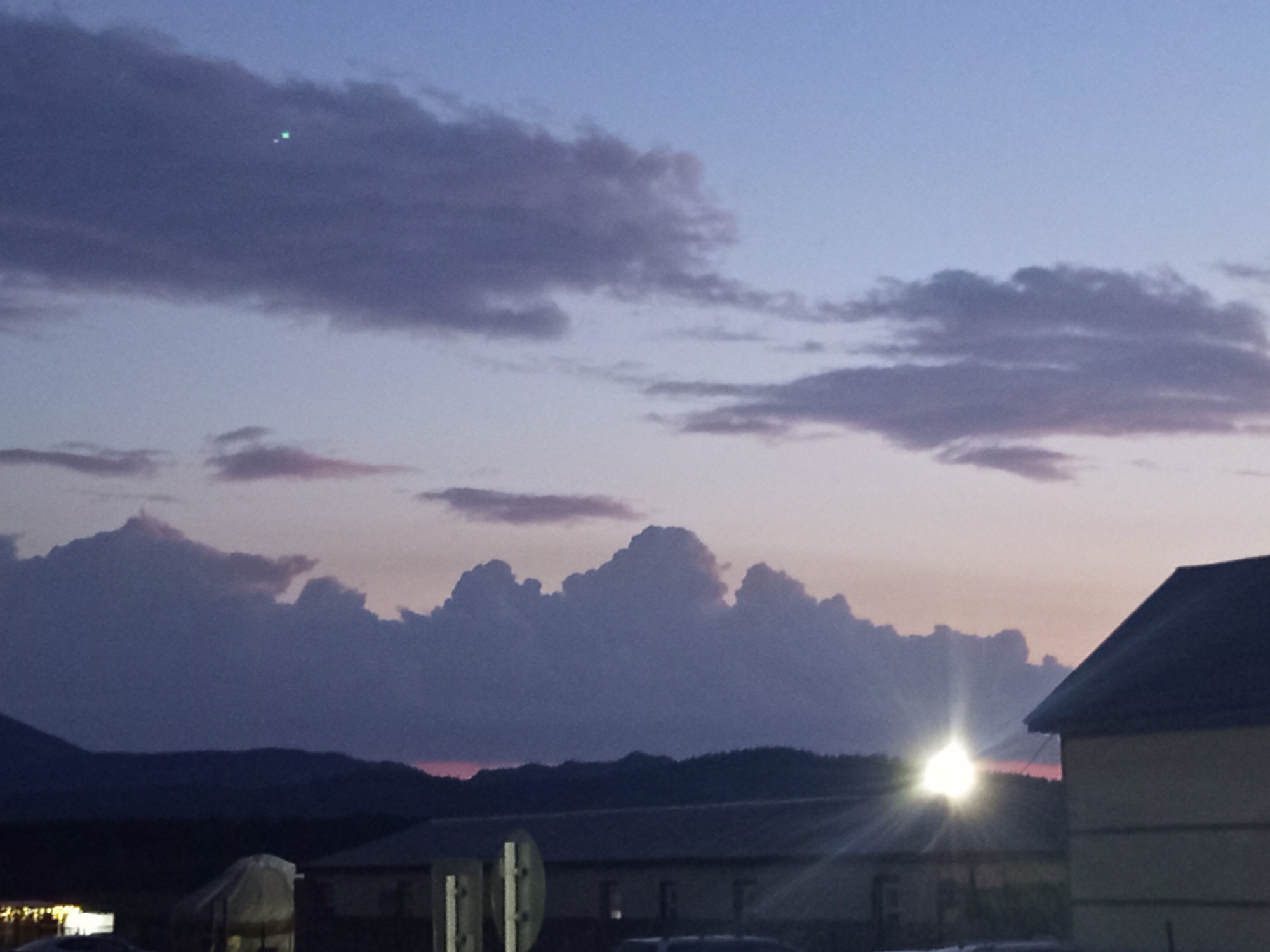 This image captures a twilight sky over the Баянауыл ауылдық округі in Kazakhstan. The sky transitions from a pale blue at the top to a softer gradient of pink and lavender near the horizon, where dramatic, bulbous clouds are silhouetted against the fading light. A small, bright green light is visible in the upper left portion of the sky, possibly a distant aircraft or star. Below the clouds, a dark, hilly landscape forms the horizon. In the foreground, the roofs of several buildings are visible, with one building on the right featuring a dark, peaked roof. A bright light source, likely a streetlamp or strong light fixture, emanates a sunburst effect from behind some structures in the mid-ground, casting light onto the surrounding buildings and landscape. The overall impression is one of a rural or semi-rural setting at dusk.