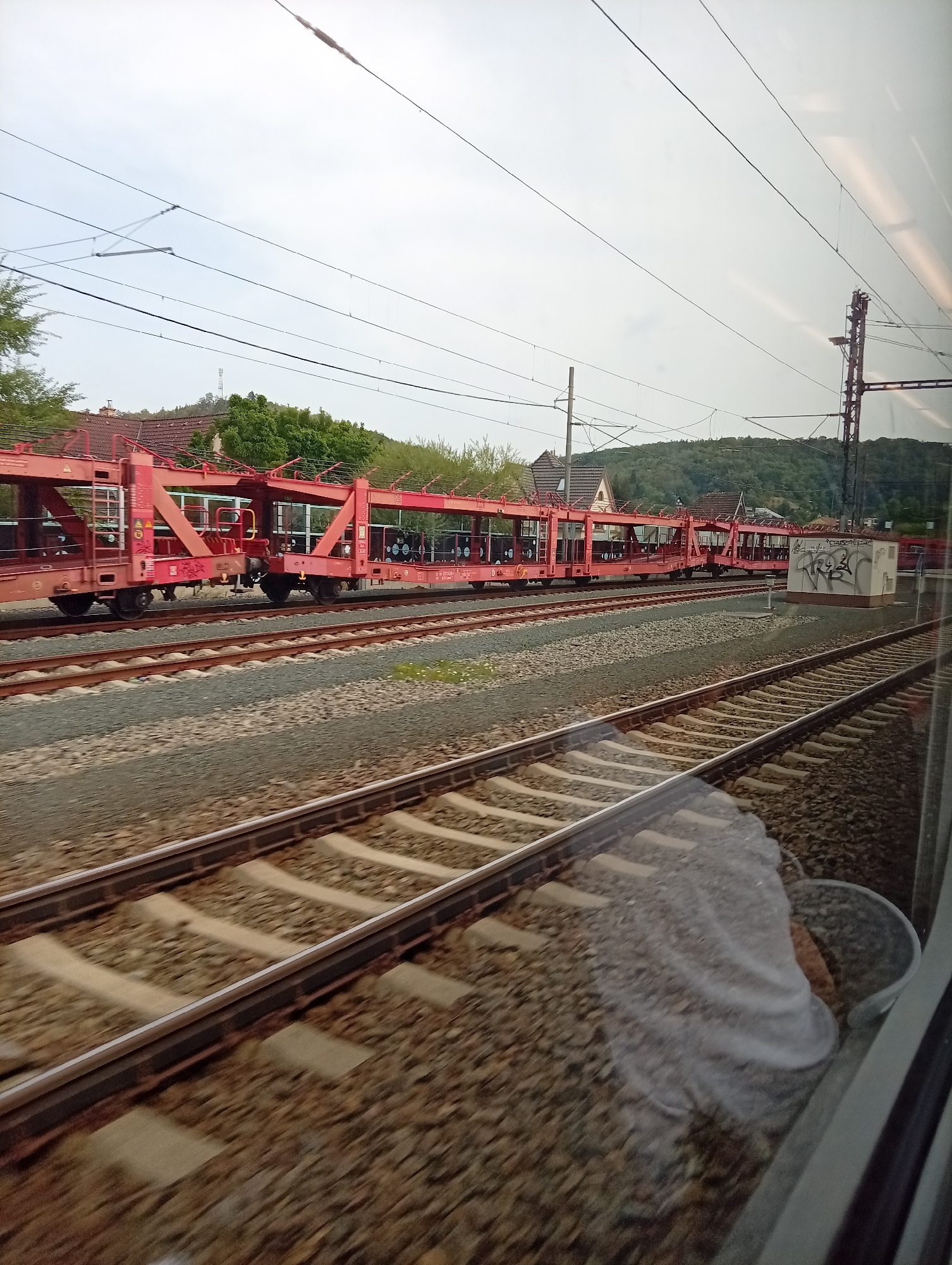 The image depicts a train with red car carriers traveling on a railway track. The train is likely moving, as indicated by the motion blur on the tracks and the surrounding environment. In the background, there are houses with red roofs, trees, and a hill covered in greenery. Overhead, there are electrical wires for the train and a utility pole. The sky is overcast. The scene appears to be in a semi-rural or suburban area. Based on information associated with the image, the location is identified as Kuřim, Czechia. There are no visible people in the frame.