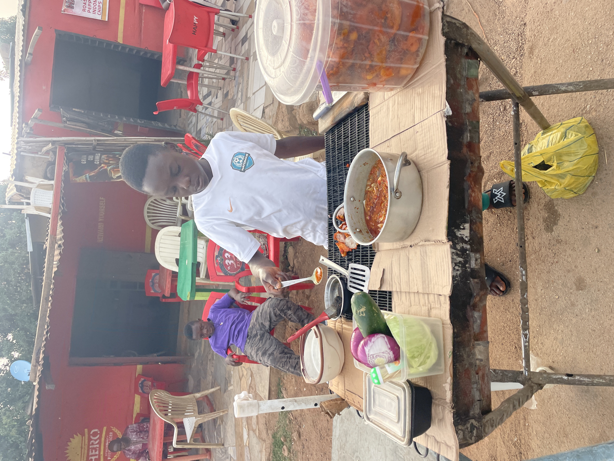 The image captures an outdoor food stall or casual eatery in Jos, Nigeria, during daylight hours. A young man, wearing a white t-shirt with a "NATOMAS" logo and a Nike swoosh, stands behind a makeshift counter fashioned from a metal frame and cardboard. He is actively engaged in preparing food, holding a large spoon and stirring contents in a silver pot placed over a grill. To his right, a large clear plastic container holds what appears to be a stew or chopped ingredients.
On the counter, fresh ingredients like a cucumber, red onions, and cabbage are stored in plastic containers, alongside cooking utensils such as a spatula and a brush. Underneath the counter, a black slipper with a white "K" logo is visible, and a yellow plastic bag hangs from the stand.
In the background, a red building with open doorways suggests a small shop or restaurant. Various red and some cream-colored plastic chairs are arranged around small tables. A man in a purple shirt and camouflage pants sits on a red chair, observing the cooking activity. The ground is unpaved, consisting of dirt or gravel.
Visible text on signs includes:
- On the red building: "HAPPY", "ROAST", "PEPPER", "INDOMIE", "CHIPS". Another sign reads "HERO", "QUALITY", "PREMIER LAGER", "REWARD YOURSELF", "GRADI", "COLD", and "GUINNESS".
- On the young man's t-shirt: "NATOMAS" and a Nike swoosh.
The scene conveys a lively, casual street food vendor setup.