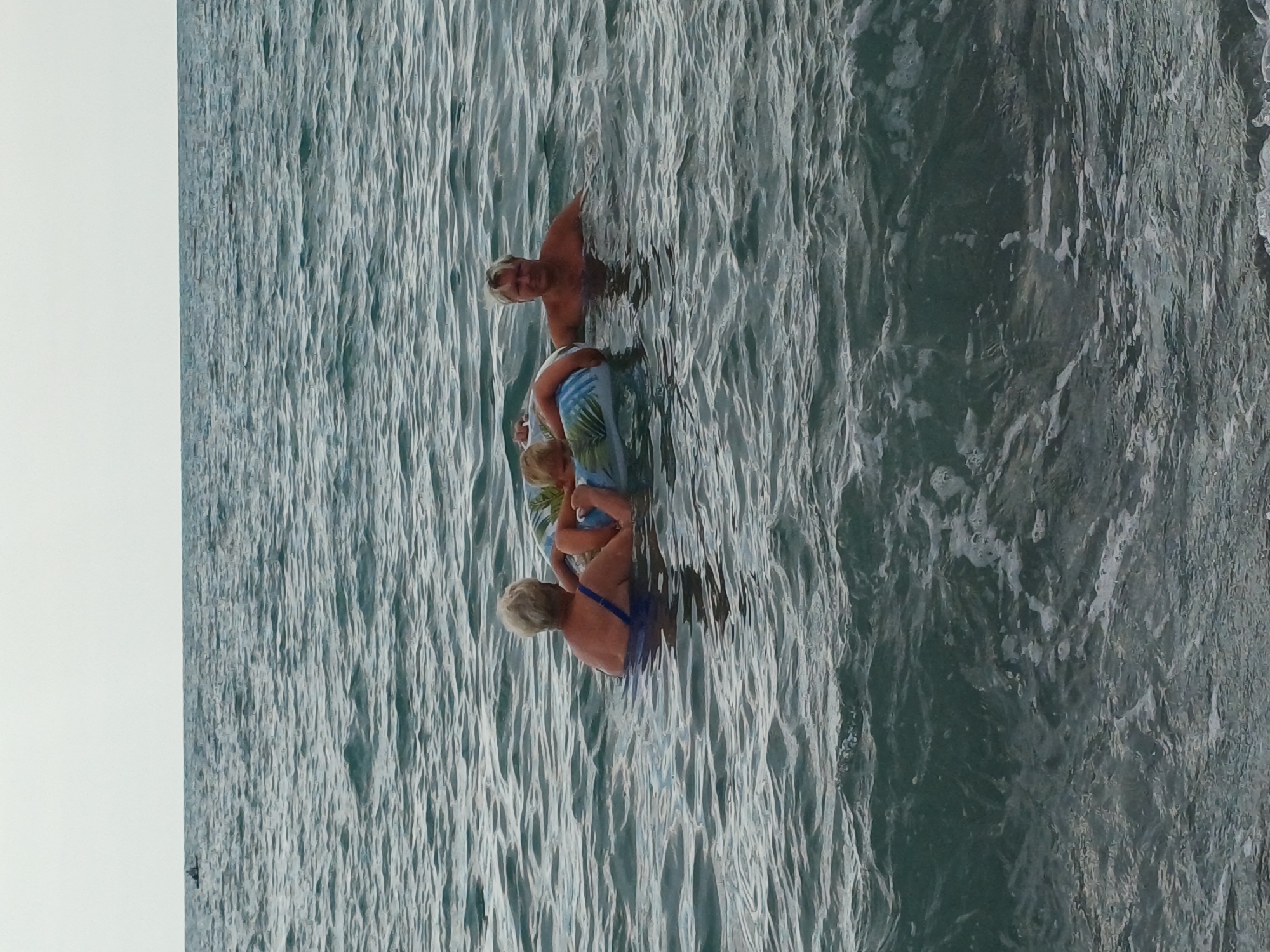 This is a medium-distance, eye-level shot of three people in the Black Sea off the coast of Sochi, Russia. The sky is overcast and the water is choppy with moderate waves.
In the foreground, the waves are breaking and frothing. In the middle ground, a woman with short blonde hair, wearing a blue swimsuit, is holding a child in an inflatable ring. The child has blonde hair and is facing away from the camera. To the right of the woman and child, a man with blonde hair and a tanned complexion, wearing no shirt, is treading water and looking towards the camera. He appears to be standing, as his chest and shoulders are visible above the water. The inflatable ring has a tropical leaf pattern.
In the far background, a small boat with a person in it is visible on the horizon. The scene suggests a leisurely day at the beach.