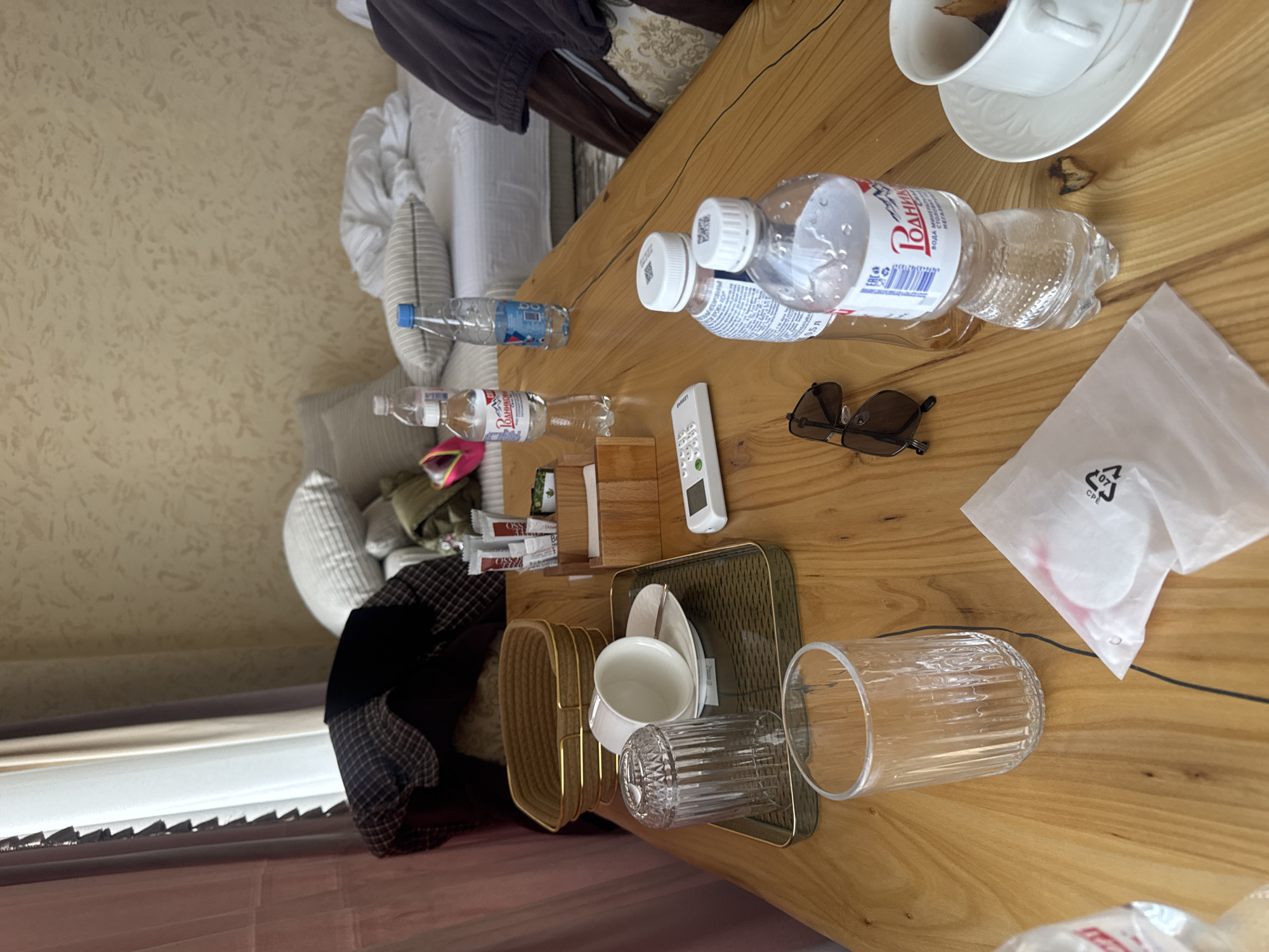 The image shows a wooden table in a room, likely a hotel room. Visible on the table are several bottles of water, one of which is labeled "РОДНИК" (Rodnik). There's also a pair of sunglasses, a white remote control, a wooden box with various small items (possibly toiletries), a golden tray holding a cup and saucer, a glass, and a white plastic bag containing what appears to be cotton pads. In the background, there's a bed with pillows and a patterned curtain near a window. Based on the label on the water bottle, the location could be Baksan, Russia.