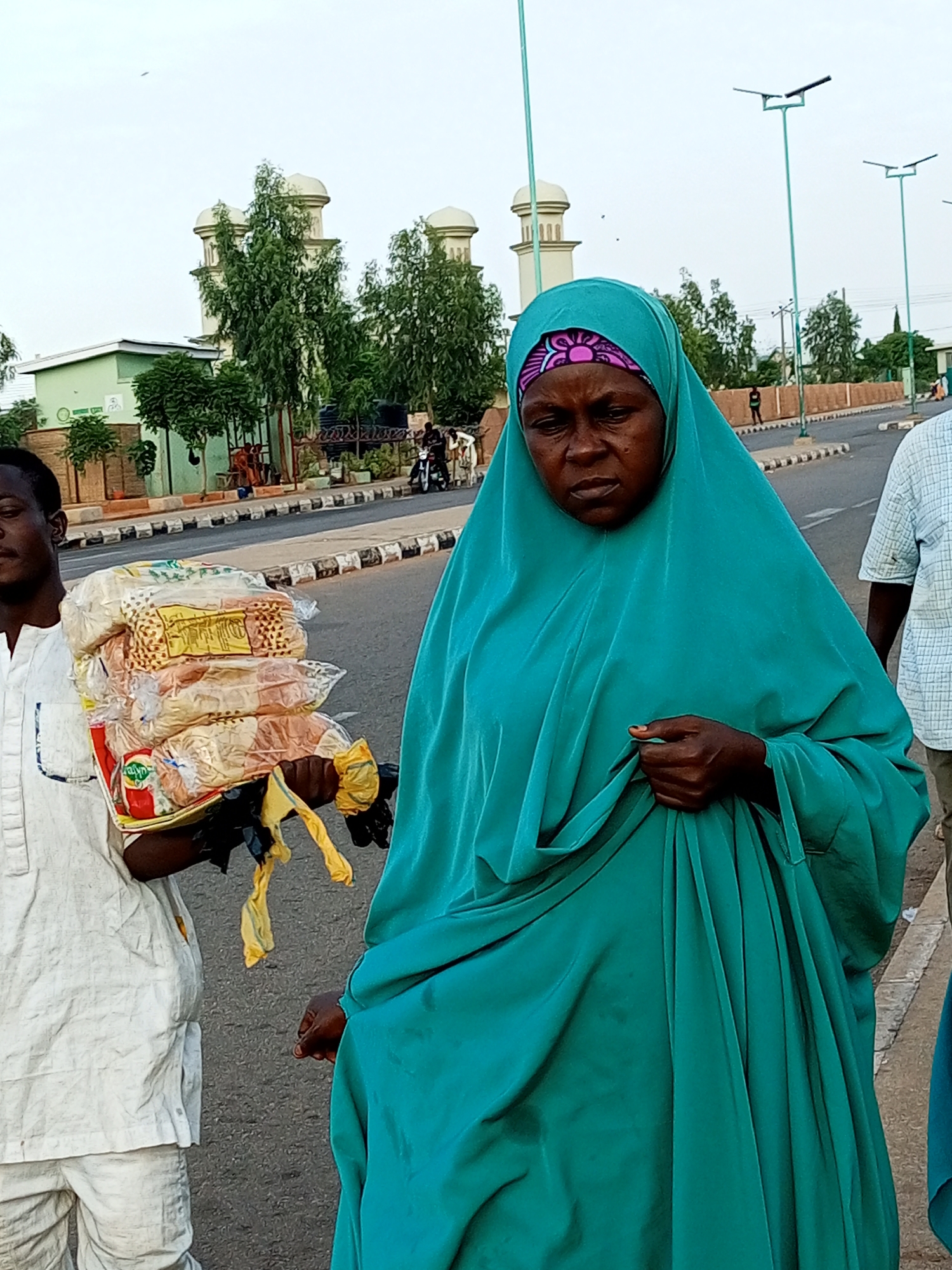 In Gombe, Nigeria, a woman wearing a teal hijab and dress is captured in a medium shot, her gaze directed downward. She stands in the foreground, her right hand holding the fabric of her dress. To her left, a person in a white outfit is partially visible, carrying a stack of bread wrapped in plastic. The background reveals a street scene with a mosque featuring twin minarets, trees, streetlights, and other individuals, some on a motorcycle, suggesting a bustling urban environment.