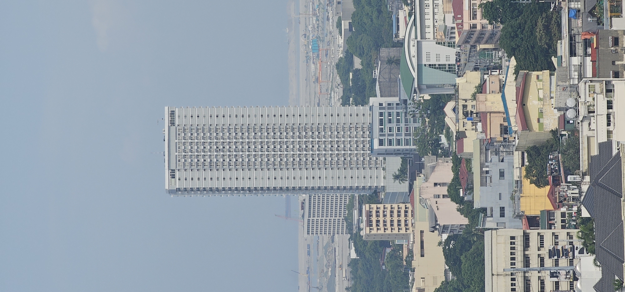 This is an aerial photograph of a dense urban landscape, likely taken from a high vantage point, showcasing a variety of buildings and infrastructure. The dominant feature is a tall, slender, white high-rise building in the center of the frame, characterized by its repetitive pattern of balconies and windows. To its left, a slightly shorter, light-colored building with a similar grid-like facade is visible.
In the foreground and middle ground, a complex network of smaller buildings of varying heights, colors, and architectural styles occupies the space. These structures range from multi-story residential or commercial buildings with beige, yellow, and light blue facades to simpler, more utilitarian structures. Greenery from trees is interspersed throughout the urban fabric, suggesting parks or tree-lined streets within the city.
In the background, a hazy, expansive area can be seen, likely representing a port or industrial zone, with cranes and shipping containers visible. The sky is a pale, overcast blue.
Given the context of the provided image and typical Makati cityscape, this photograph likely depicts an area within Makati City, Philippines, known for its high-rise developments and dense urban environment. The scene is devoid of discernible people or specific actions, presenting a static, architectural overview of the city.