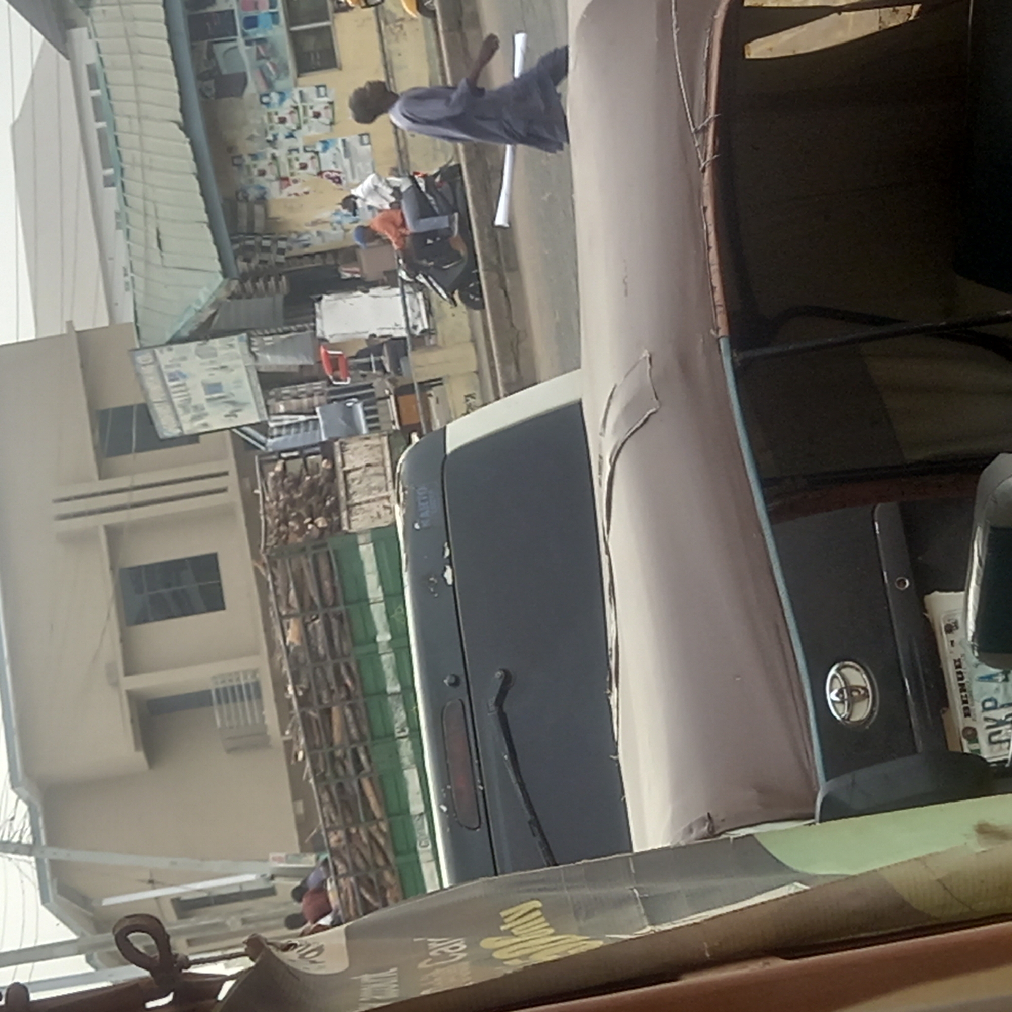 The image captures a street scene, taken from within a vehicle, likely an auto-rickshaw. The perspective is angled, looking out onto a bustling environment.
In the foreground, the upper portion of the vehicle's interior is visible, featuring a light-colored fabric canopy and a dark dashboard. A silver Toyota emblem is clearly seen on the dashboard. A Nigerian license plate is partially visible, displaying "BENUE" at the top and "CKP 4" below it. Fragmented, blurred text, possibly reading "Amunmula Car," appears on a surface in the immediate foreground.
The midground reveals a paved street or road. A person, dressed in a long, dark blue or grey traditional garment, walks away from the camera. Two white cylindrical objects are on the ground near this individual. Slightly further back, another person is seated on a motorcycle or scooter.
The background is comprised of buildings and commercial activity. On the left, a light-colored, multi-story building with multiple windows and distinctive horizontal accents is prominent. To the right, behind the street, are various structures resembling shops or market stalls. Displays of goods and several posters adorn their walls. Stacks of wooden crates or boxes are arranged in front of some of these establishments.
The scene unfolds during daytime under clear weather conditions, indicating typical daily life. The "BENUE" text on the license plate places the location in Benue State, Nigeria. No specific emotions are discernible from the image.