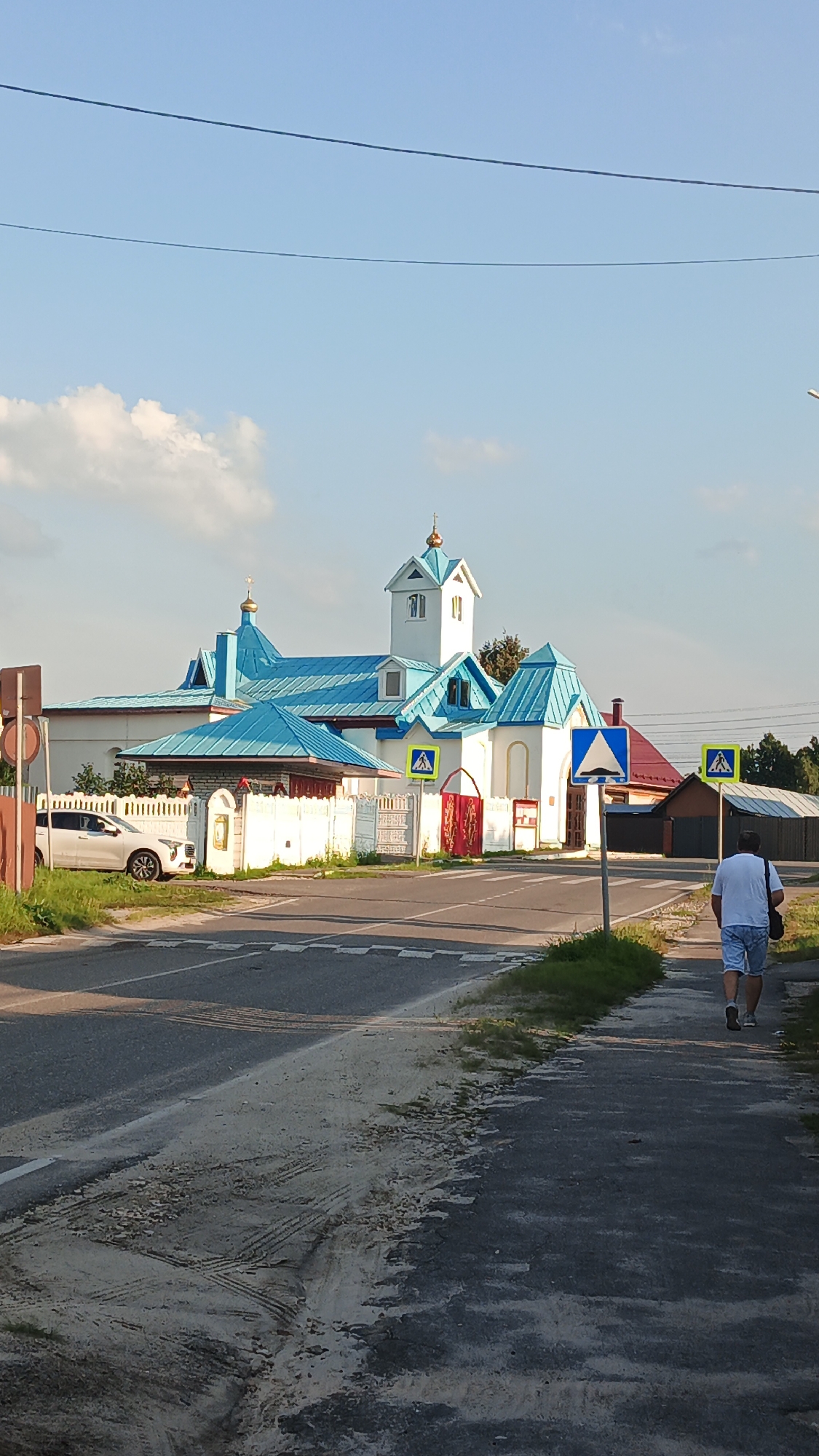In Drezna, Russia, on a clear day with a few clouds, a person walks on a sidewalk next to a road. The person is wearing a white t-shirt and light blue shorts and carrying a dark bag over their right shoulder. Ahead of them, a white building with a bright blue roof, possibly a church, stands behind a white fence. The building has several spires topped with golden crosses. A white car is parked to the left of the building, behind the fence. Several pedestrian crossing signs are visible near the road. The road itself has white markings and a speed bump. The environment is rural or suburban, with some trees and other buildings in the background.