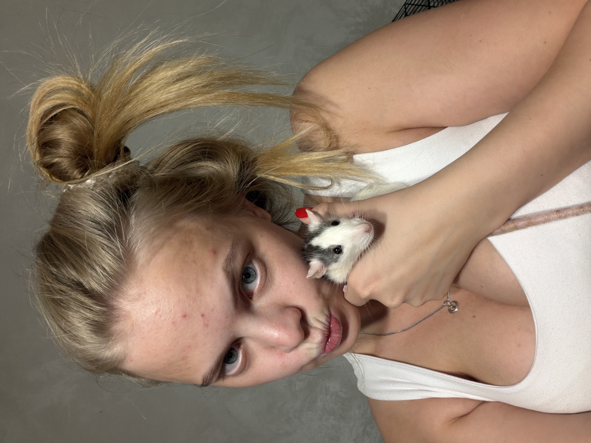 A young woman with light blonde hair styled in a messy bun and some strands pulled forward poses with a white and black hooded rat. She is wearing a white tank top and a silver heart-shaped pendant necklace. The background appears to be a plain, light grey wall. The woman is holding the rat close to her face. The location in the city of Chaikovsky, Russia is not discernable from the image.