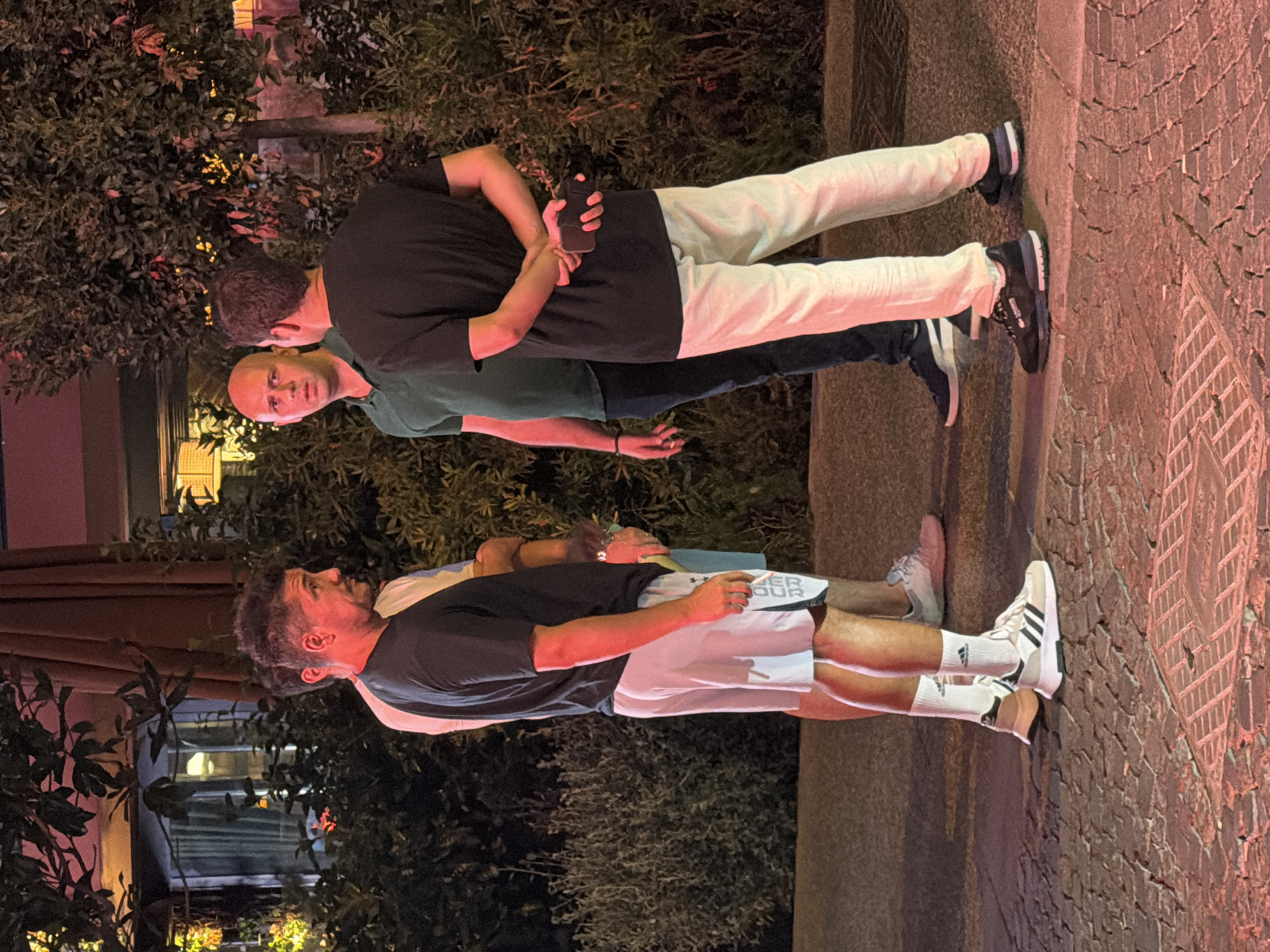 Four men are standing on a brick-paved walkway at night. The man on the far left is wearing a black t-shirt and white shorts with black stripes and lettering. He has white socks and white and black sneakers. The man next to him is wearing a light-colored t-shirt and light-colored shorts. The man in the center is wearing a dark green polo shirt and dark pants. The man on the far right is wearing a black t-shirt and light-colored pants. He has his hands clasped behind his back, holding a black cell phone. The men are facing each other and appear to be in conversation. Behind them are dense green bushes and trees, with some red and yellow lights visible in the background. The scene is lit by ambient outdoor lighting, casting reddish and yellow tones on the ground and the men. The location is identified as Kadıköy, Turkey.