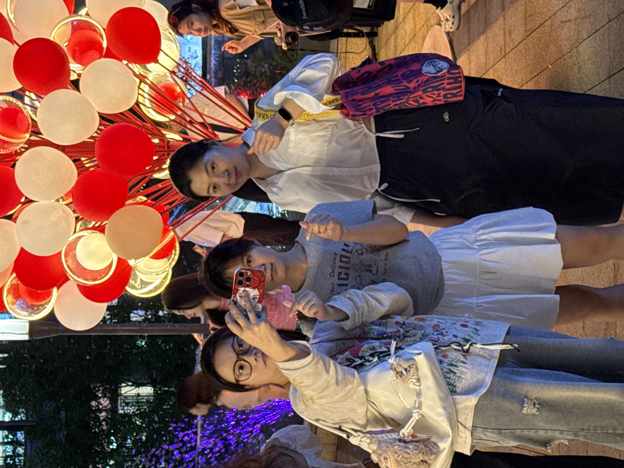 In Yuzhong District, China, a group of women are interacting with each other and taking photos with their cell phones near a display of red and white balloons. One woman is holding her phone up to take a selfie with another woman, while another woman poses with a thumbs-up gesture. The women are wearing casual clothing. In the background, there are trees and buildings illuminated with purple and blue lights.