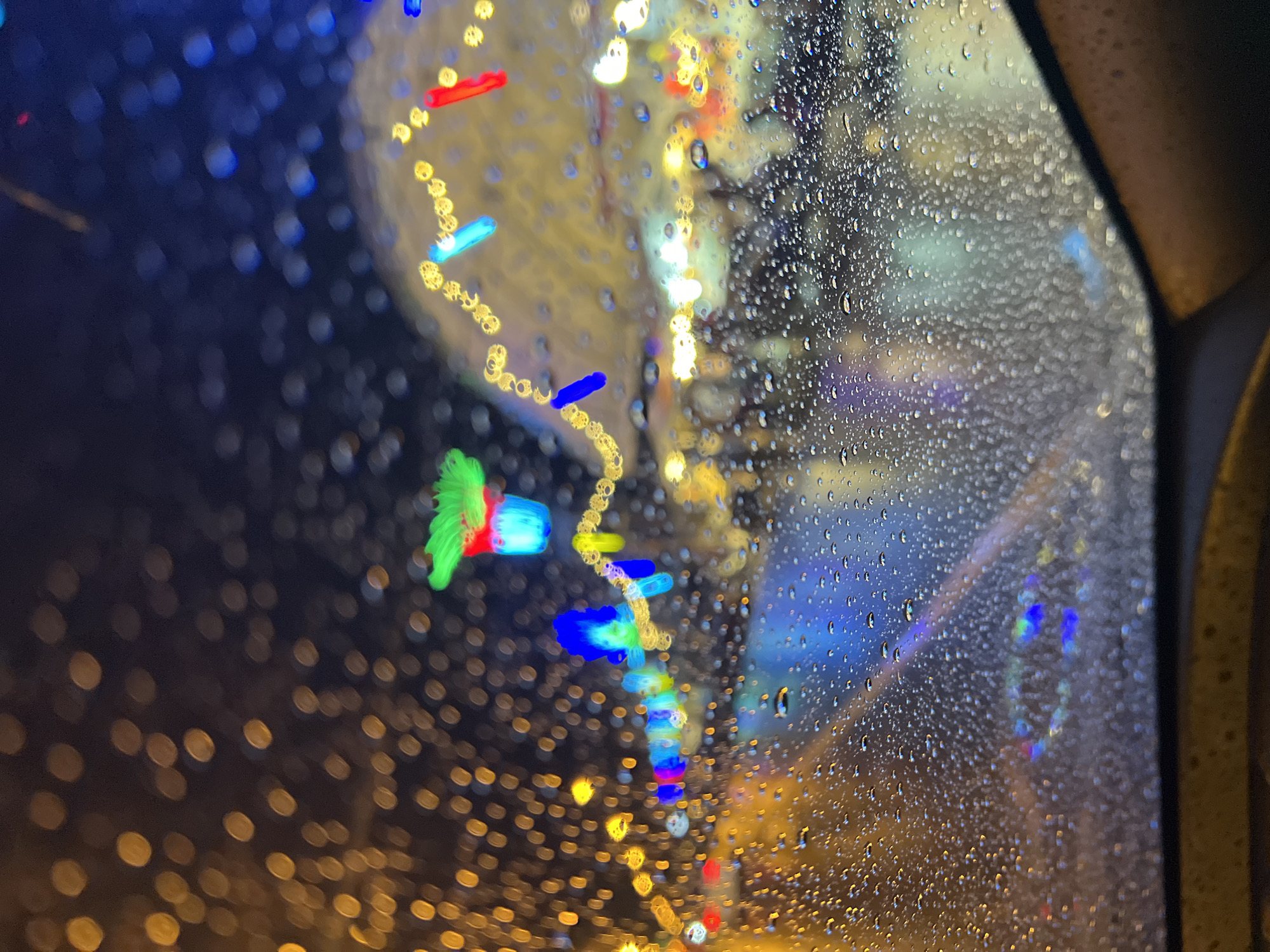 A blurred view through a rain-streaked window shows colorful lights and a dark background, possibly at night. The presence of a car frame on the right suggests the view is from inside a vehicle. The lights are out of focus, appearing as bokeh shapes of yellow, red, green and blue. It is impossible to specifically confirm the image's location in Baan Bo Phut, Thailand without additional context.