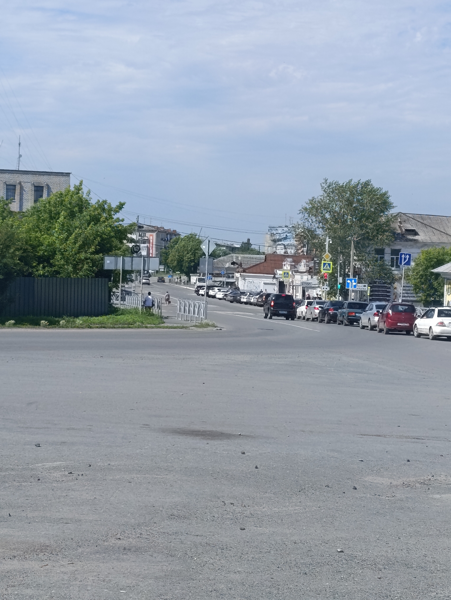 This is a daytime, eye-level, exterior shot of a street in Talitsa, Russia, likely on a cloudy day given the overcast sky. The foreground is dominated by a grey asphalt road, with scattered small rocks and debris visible on its surface. In the middle ground, there is a wide asphalt intersection or parking area. To the left, a dark grey metal fence borders a patch of green grass.
Beyond the fence, a line of cars is parked or stopped along the side of a road that recedes into the distance. The cars are of various colors including white, black, silver, and red. A few pedestrians are visible near the road. One person in a white shirt and dark pants is seated on a bench, and another person in a light-colored top and dark pants is walking. Further down the road, more cars and buildings are visible, including a multi-story brick building on the left and other structures with pitched roofs on the right. Several traffic signs are present, including pedestrian crossing signs with yellow triangles, a traffic light, and directional signs. Trees with green foliage are scattered throughout the scene. The background features a light blue sky with scattered clouds. The scene suggests a typical urban or suburban street environment with moderate vehicular and pedestrian activity.