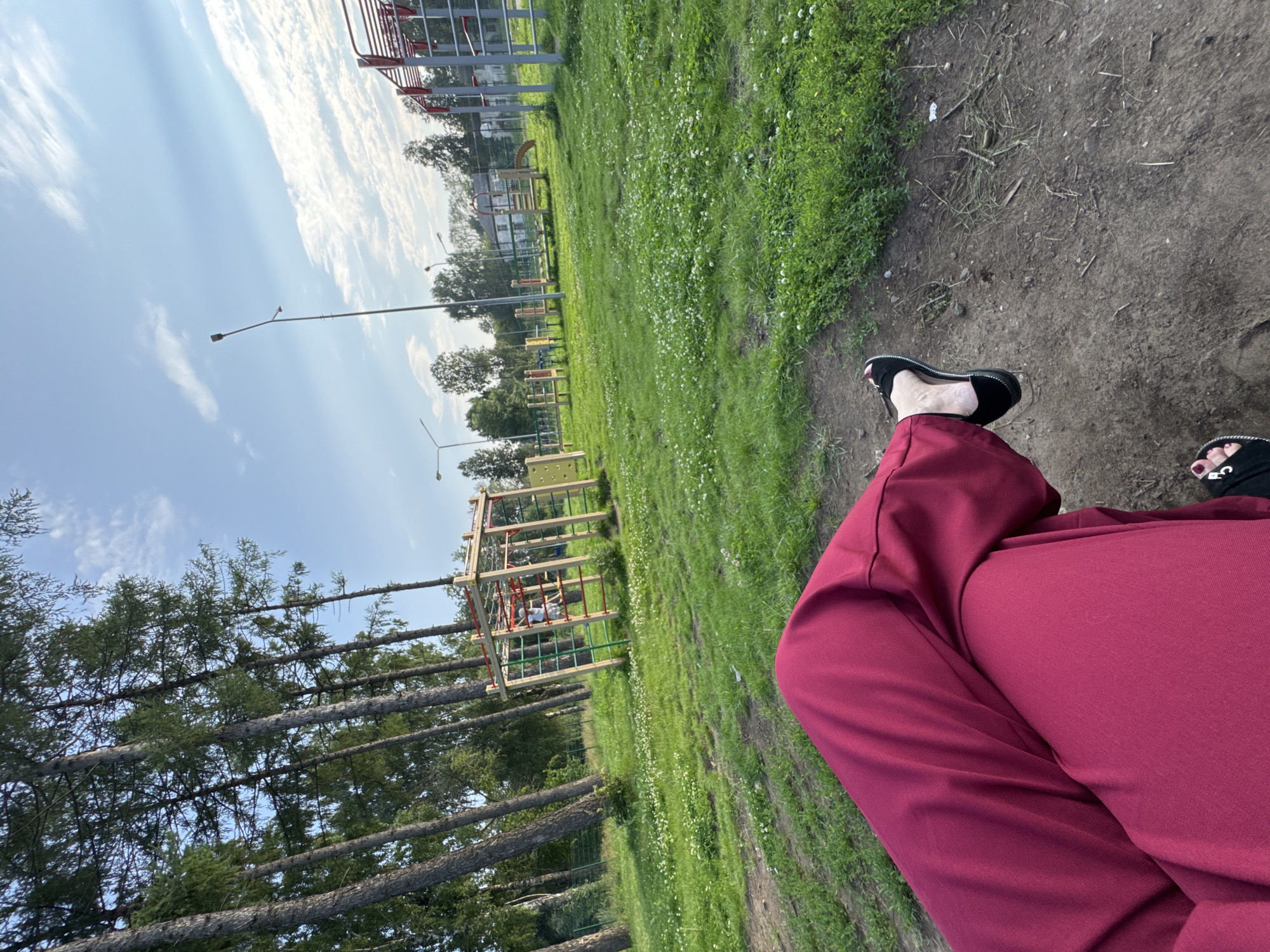 The image depicts an outdoor scene in Chaikovsky, Russia. The viewpoint is from a person sitting on the ground, with their legs, clad in burgundy trousers, and feet, wearing black sandals, prominently in the foreground. The setting appears to be a park or recreational area with a large grassy field dotted with small white flowers. In the background, there are tall evergreen trees, a playground structure with orange and green bars, and what looks like outdoor exercise equipment. Lampposts are visible throughout the park. The sky is blue with scattered white clouds, suggesting it is daytime with fair weather. The overall impression is one of leisure and relaxation in a natural, communal outdoor space. There is no visible text in the image.