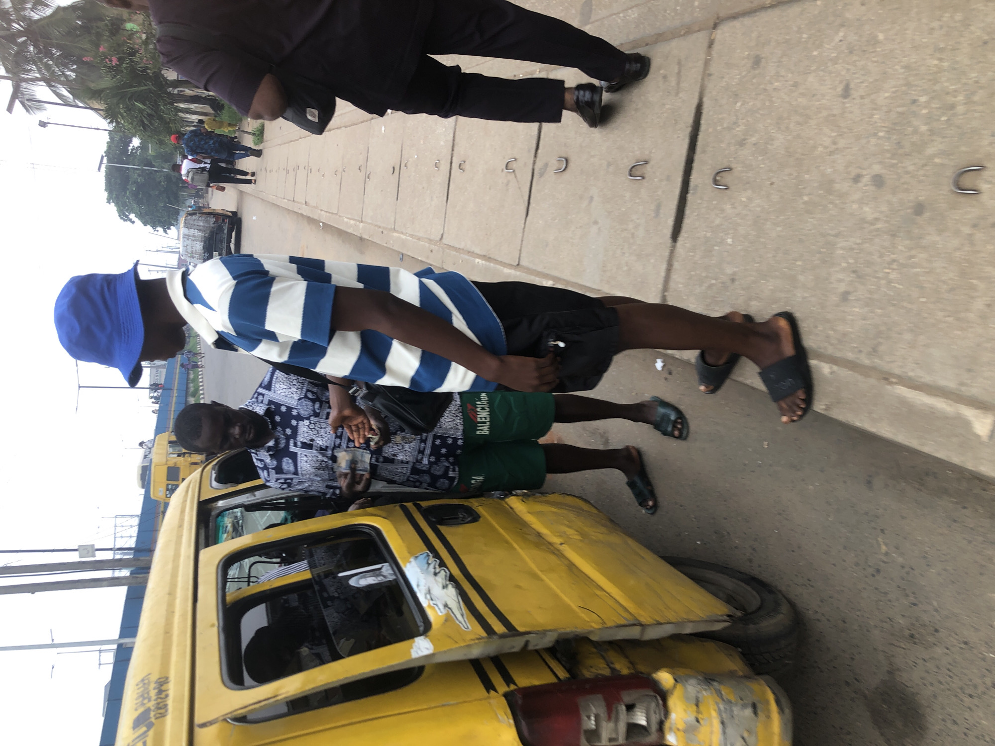 In Ikeja, Nigeria, two men are standing by an open yellow taxi van. One man, wearing a blue bucket hat and a blue and white striped shirt with black shorts, is facing away from the van. The other man, dressed in a patterned shirt and green shorts with "BALENCIAGA" written on them, is standing next to the van, holding what appears to be money in his hands. The taxi van has an open side door, revealing a dark interior. The scene takes place on a paved road next to a sidewalk with concrete slabs. In the background, other vehicles and pedestrians can be seen, along with trees and overhead structures, indicating an urban environment.
