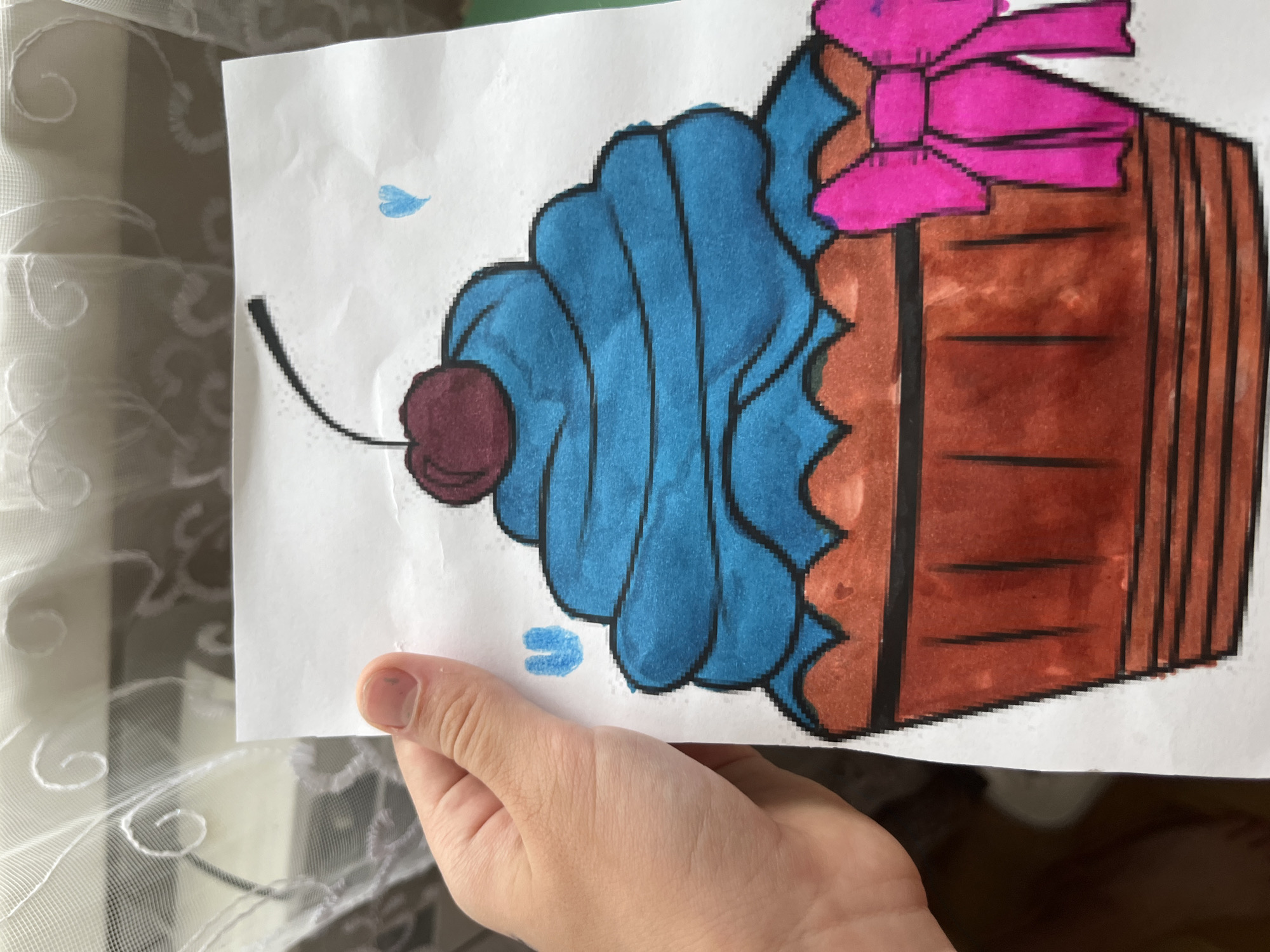 The image displays a child's drawing of a cupcake, held by a person's hand. The drawing features a blue frosting topped with a red cherry, a brown cupcake liner, and a pink bow. There are small blue heart shapes near the cupcake. The background consists of sheer white curtains. There is no visible information or context to suggest a location in Armavir, Russia.