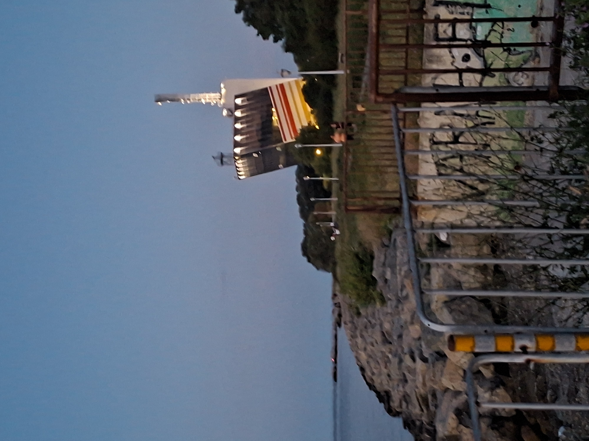 The image depicts a view from a rocky shoreline looking towards a modern, uniquely shaped building illuminated at dusk. The building, which appears to be a large structure with a leaning facade and a tower, has multiple levels and is decorated with colorful horizontal stripes in red, white, and yellow on one side. The sky is a deep blue, transitioning to a lighter hue near the horizon, suggesting twilight. In the foreground, a metal fence with graffiti on a wall behind it partially obstructs the view. There are some people visible in the middle ground near the building, appearing to be standing or sitting. The environment includes a body of water on the left, likely the Black Sea, and trees and vegetation surrounding the building and shoreline. The overall scene suggests a public space or park area in Varna, Bulgaria, possibly near a waterfront attraction or landmark.