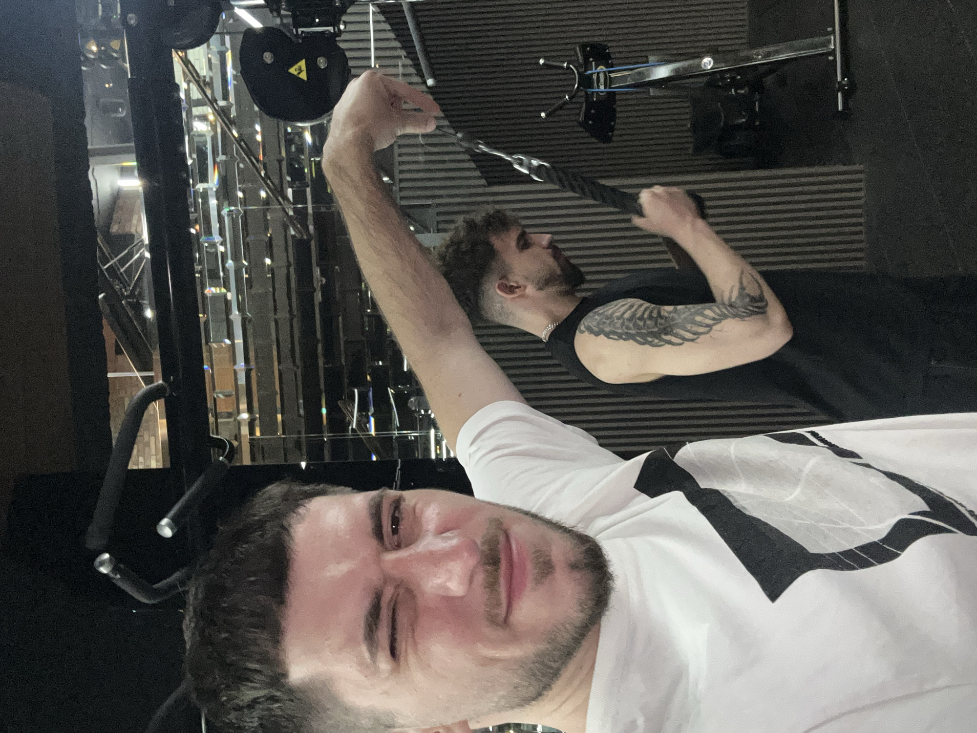 Two men are in a gym. The man in the foreground is taking a selfie while winking. He is wearing a white t-shirt with a geometric design. Behind him, a second man is pulling on a cable machine. He is wearing a black tank top and has a tattoo on his arm. The gym has mirrored walls and modern equipment. The specific location within Sofia, Bulgaria cannot be determined from the image.
