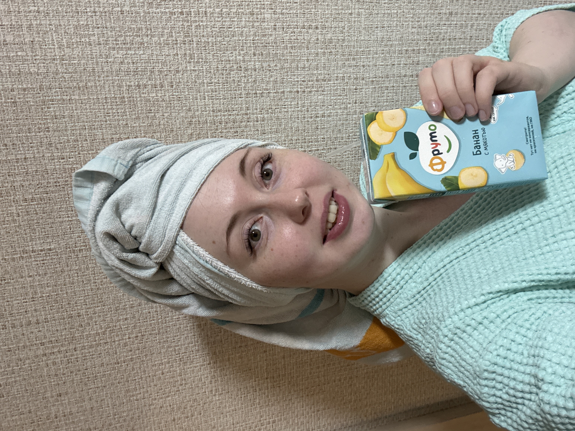 A young person, likely female, is depicted from the chest up, lying down and smiling gently at the camera. Their light brown hair is wrapped in a light blue and white striped towel, and they are wearing a light blue waffle-knit robe. They hold a small, light blue juice box in their right hand. The juice box features illustrations of bananas and banana slices and displays the brand "Фруто" (Fruto). The product is identified as "Банан с мякотью" (Banana with pulp). Additional text on the box includes "как сочног" (how juicy) and a claim stating, "Смотрите! Тут нет ни консервантов, ни красителей, ни ГМО" (Look! There are no preservatives, no dyes, no GMOs). The background is a textured, light beige wall covering, suggesting an indoor, casual setting, possibly at home after a shower. The person's expression is relaxed and friendly.