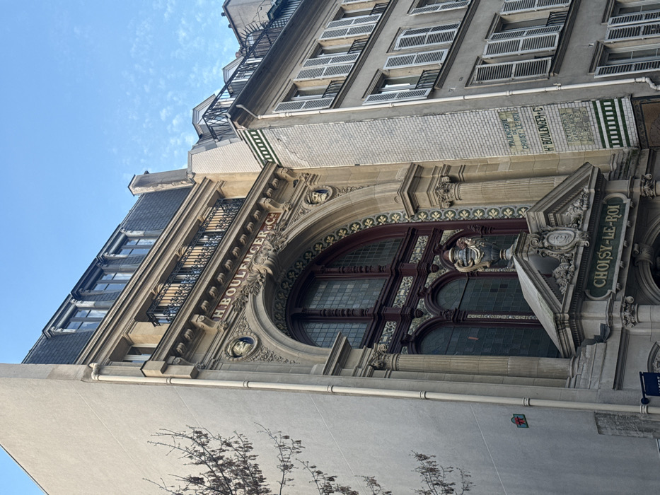 The image displays the ornate stone facade of a historic building located at 79, Rue de la Boétie, 75008 Paris, France, under a clear blue sky. The central structure features a large arched window with dark-framed, multi-paneled leaded glass, flanked by decorative pilasters and intricate ceramic tiling in green, gold, and white patterns. Above this arch, the inscription "H. BOULENGER & C°" is carved, positioned between two circular medallions each containing a sculpted male bust. Below the main window, a prominent stone pediment is visible, adorned with intricate carvings and topped by a decorative urn. The inscription "CHOISY-LE-ROI" is present on the pediment's lower section. Above the main arch, the building features a floor with balconies protected by wrought-iron railings, culminating in a dark mansard roof with multiple dormer windows. To the right, an adjacent building, typical of Parisian Haussmannian architecture, displays a lighter-toned facade with several floors of windows fitted with closed white louvered shutters. A vertical tiled sign on the wall connecting the two buildings advertises "FAIENCERIE DE CHOISY-LE-ROI," "H. BOULENGER & C°," and "REVETEMENTS CARRELAGES MOSAIQUES" using green and white ceramic tiles. To the left, a plain, lighter-colored wall is visible with a section of bare tree branches extending into the frame. No individuals or specific activities are depicted.