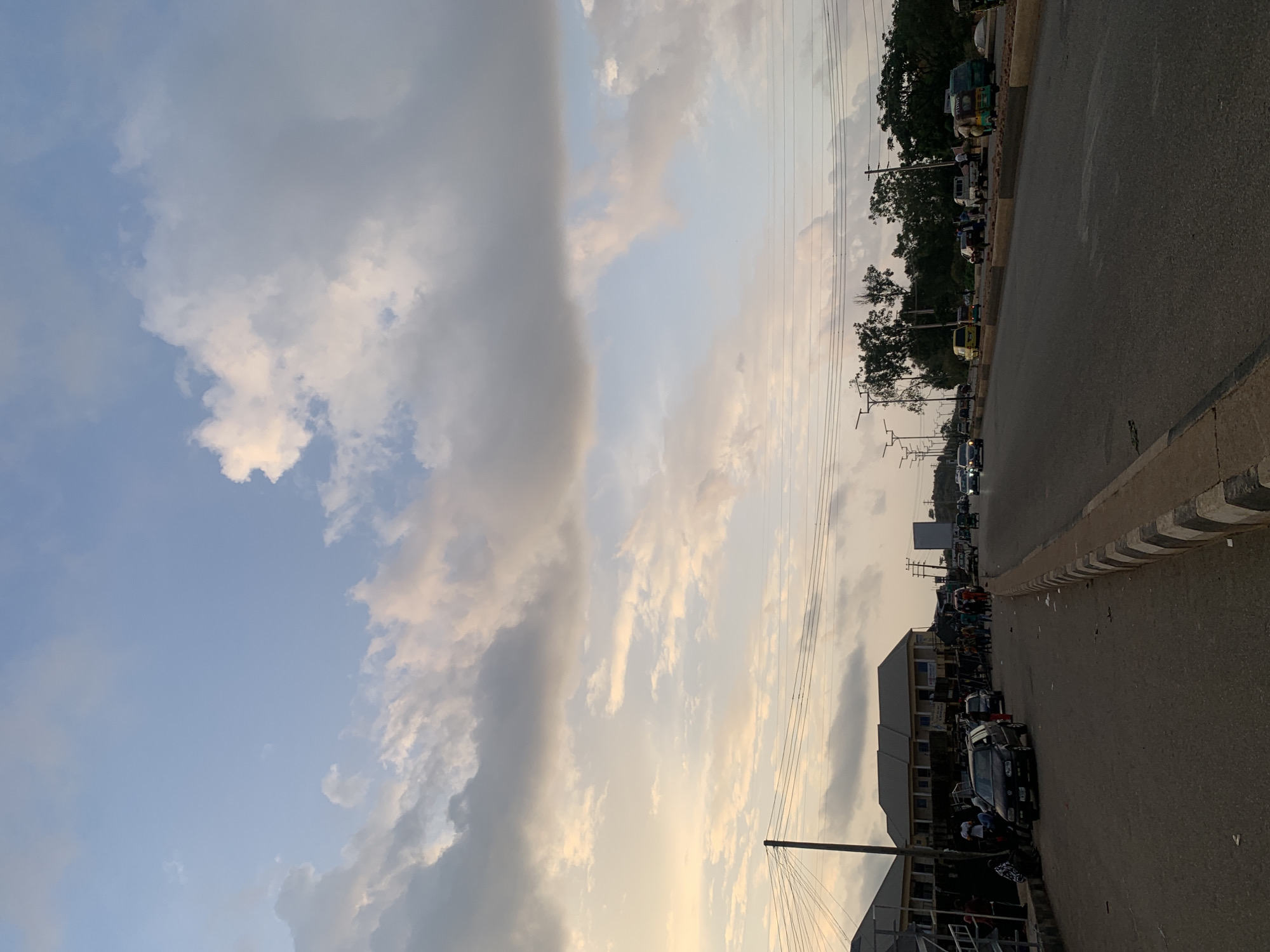 The image shows a street scene in Gura Top, Nigeria. The sky is visible with various cloud formations. The street has traffic, including cars and auto rickshaws ("Keke Napep"). Buildings line the street, and people are visible on the side of the road. Trees are also visible along the roadside. Electrical wires span the area. The lighting suggests it might be late afternoon or early evening.