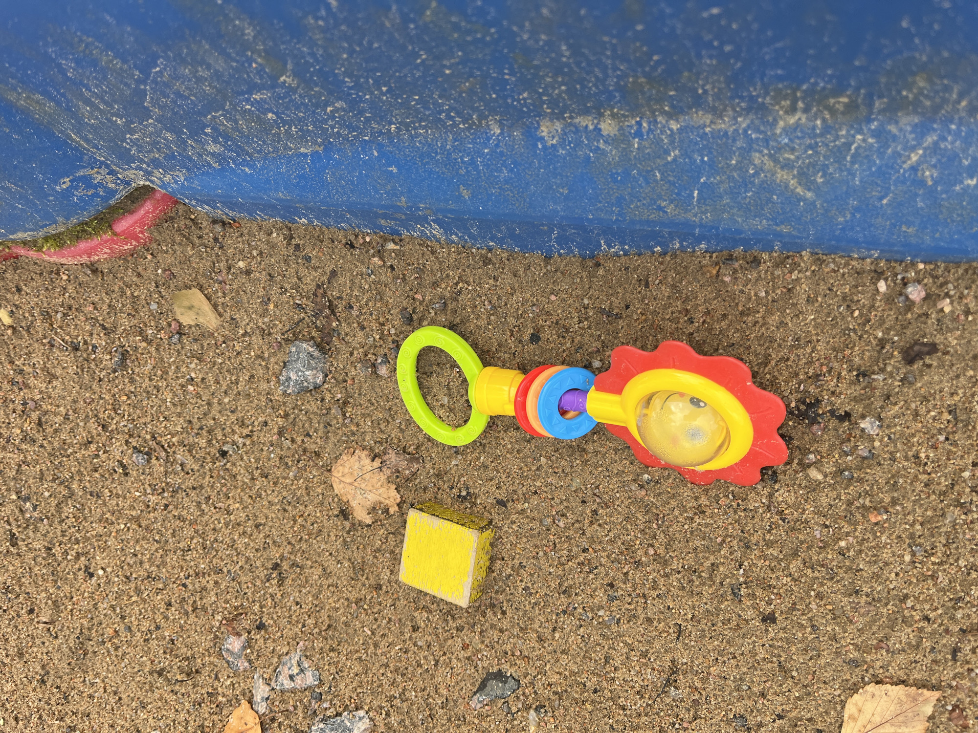 The image shows a close-up, eye-level view of a sandy area, likely a playground, with a blue plastic structure on the right side. The sand is a light brown color and contains small pebbles and a few fallen leaves. In the center of the frame, a colorful baby rattle is lying on the sand. The rattle has a green loop at the top, followed by red, blue, and purple rings. At the bottom is a red flower-shaped piece with a yellow center that contains a clear plastic ball with a yellow object inside. To the left of the rattle, a small yellow wooden block is also present. The blue structure appears to be a piece of playground equipment, possibly a slide or a climbing frame, and it has a textured surface with some white dust or debris. The lighting suggests it is daytime. The scene is devoid of any human presence. This image was taken in Il'ino, Russia.