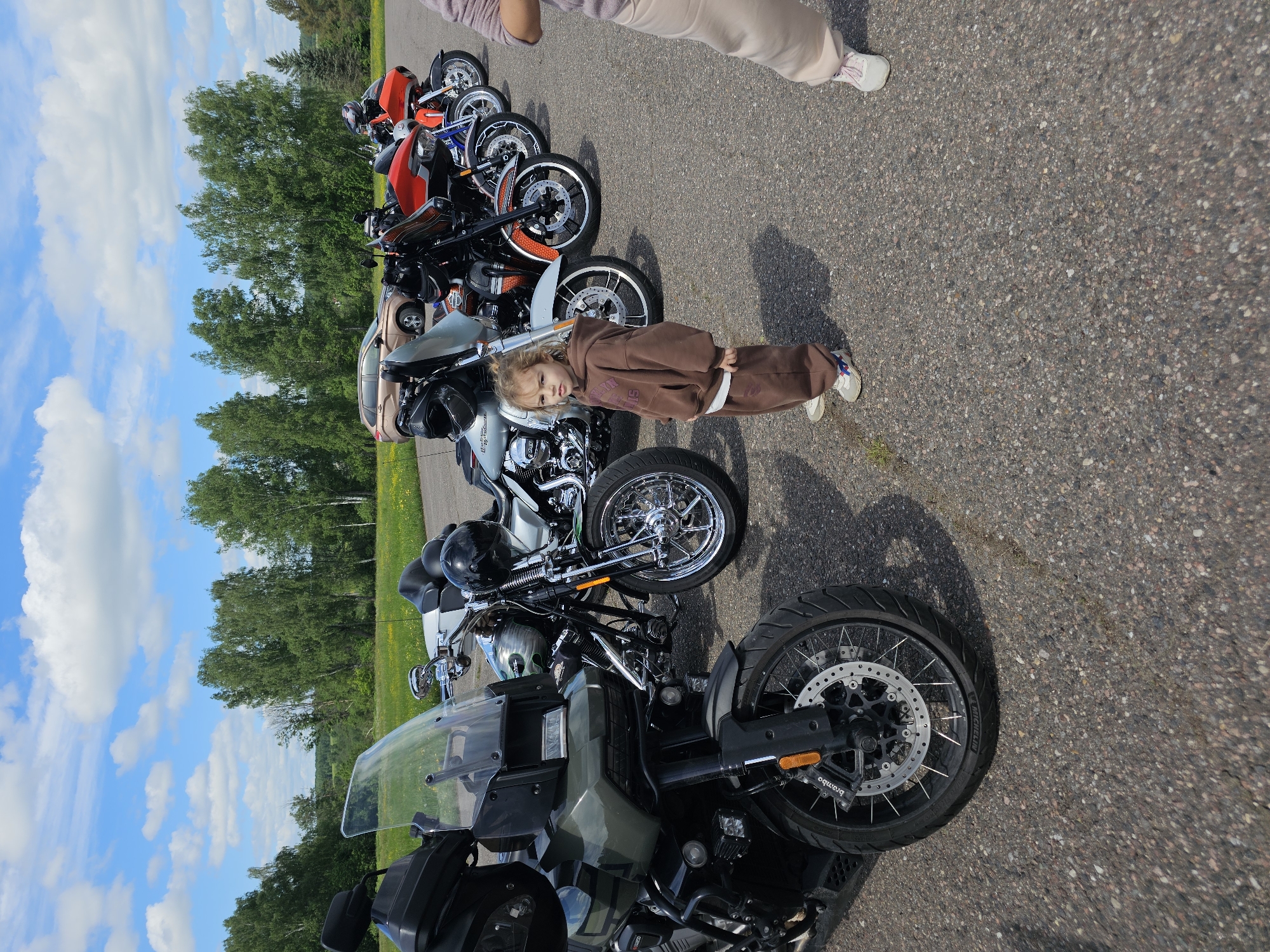 A line of motorcycles is parked on an asphalt surface next to a grassy area with trees. A young child wearing a brown sweatsuit stands in front of the motorcycles. A woman's legs are visible at the top of the image. The location is Borodinskogo Muzeya, Russia.