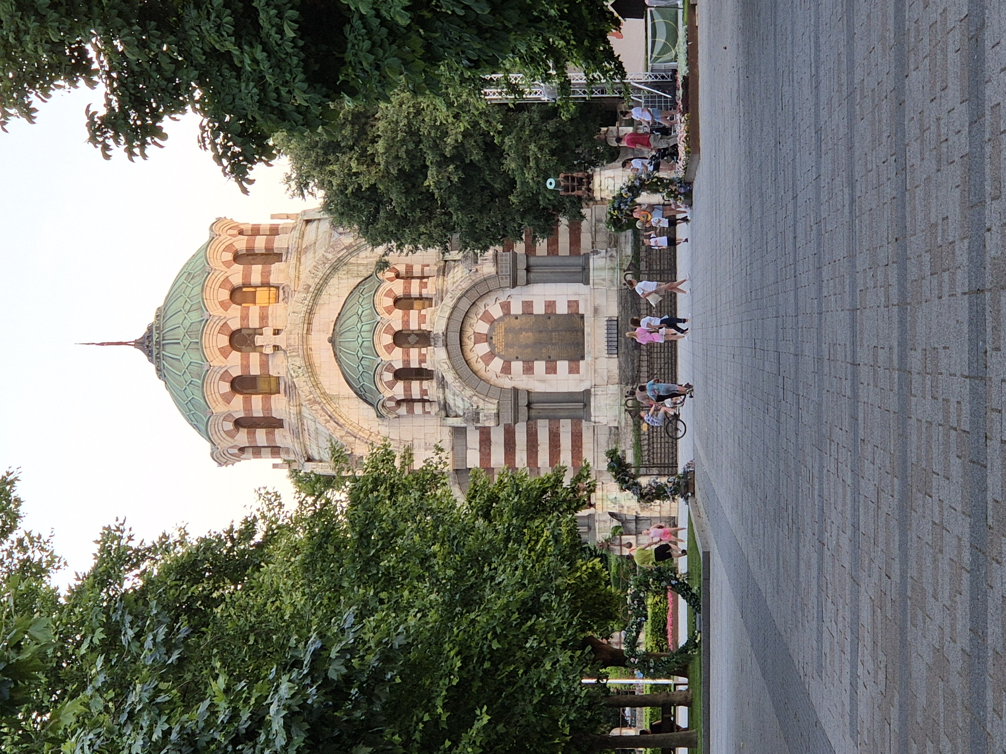 The image shows a wide-angle, eye-level view of a paved plaza in front of a large, ornate building with multiple domes, likely a church. The building features a facade of alternating red and white bands, with arched windows and decorative stonework. Green trees with lush foliage flank the building on both sides, framing the central structure.
In the foreground, a wide paved walkway stretches towards the building. Several people are present in the plaza. To the left, a man in a green shirt and dark shorts is standing near a decorative floral arch. To the right, a group of people are gathered near a stage setup. Some are walking, while others are standing. A bicycle and two scooters are visible near this group, with at least one child on the scooter.
The sky is a pale, overcast hue, suggesting either early morning or late afternoon light. The overall atmosphere appears to be casual and public, with people enjoying the space. The scene is dominated by the grandeur of the architecture and the vibrant green of the surrounding trees.