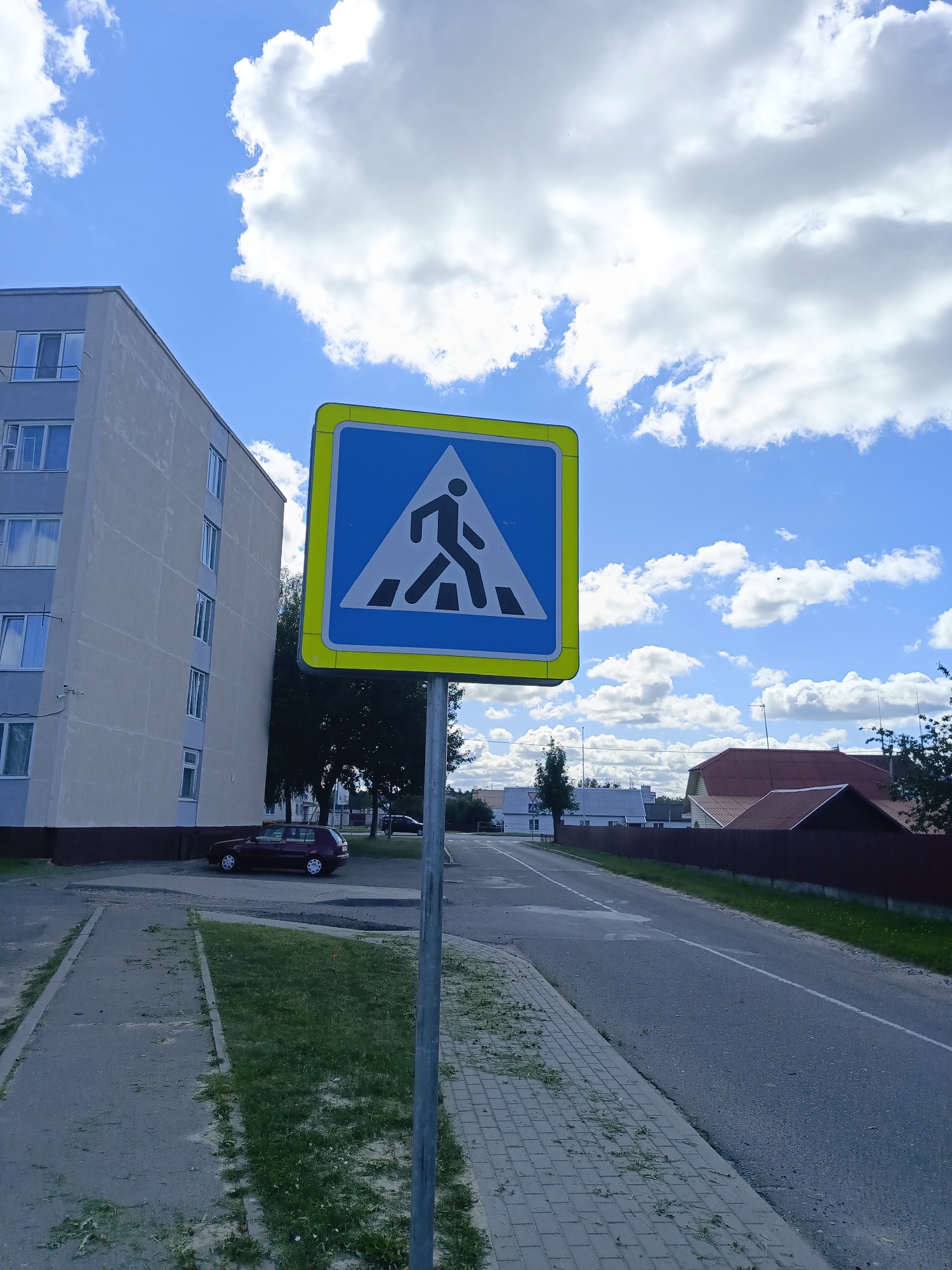 An eye-level, medium shot captures a pedestrian crossing sign on a metal pole on a bright, cloudy day in Ivatsevichy, Belarus. The sign is a blue square with a yellow border, depicting a black silhouette of a person walking on a crosswalk in white. To the left of the sign, a tall, light-colored apartment building with multiple windows runs vertically. In front of the building, a small, dark red car is parked. A paved sidewalk runs parallel to the street in the foreground, bordered by a strip of grass. The street curves to the right, and across the street, several houses with red roofs are visible behind a wooden fence. Trees and more houses are spread across the horizon under a blue sky with scattered white clouds.