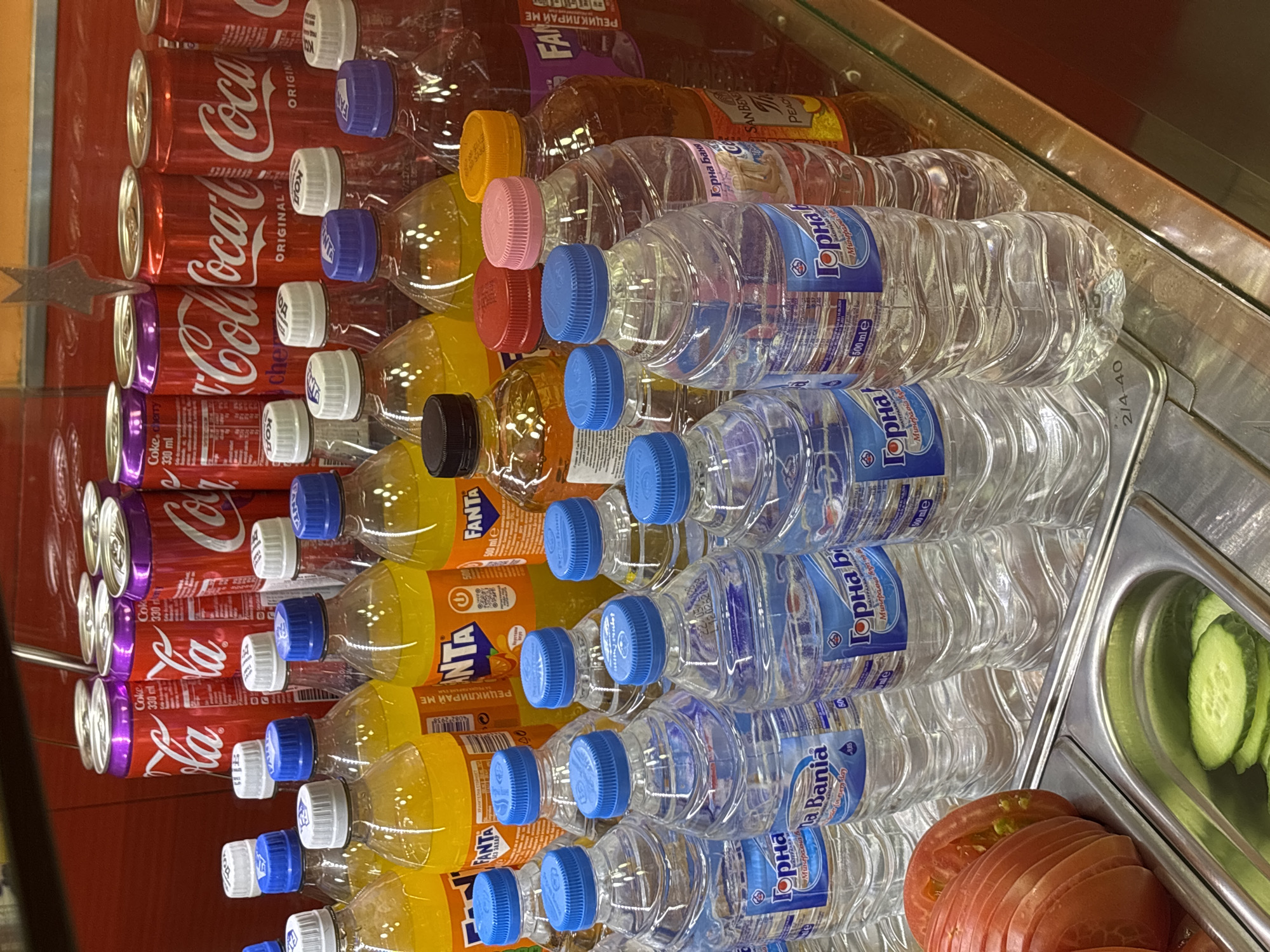 The image shows a display of beverages and sliced vegetables in a refrigerated case. In the background, rows of canned Coca-Cola are visible, some with purple accents. In front of the cans are several bottles of Fanta and other soft drinks with orange and yellow liquids. In the foreground, there are numerous plastic bottles of clear water with blue caps, bearing labels in Cyrillic that read "Горна Баня" (Gorna Bania), a brand of mineral water originating from Bulgaria. To the left, sliced tomatoes and cucumbers are presented in metal compartments, suggesting a food service setting, possibly a buffet or a self-service salad bar. The overall environment appears to be indoors, with a reflective surface likely representing the glass of the refrigerated display. The presence of Bulgarian text on the water bottles indicates the location is likely in Bulgaria, and given the specific brand of mineral water and the general appearance of the retail environment, a probable location within the city of Sofia, Bulgaria. There are no people visible in the image, and no actions or interactions are depicted.