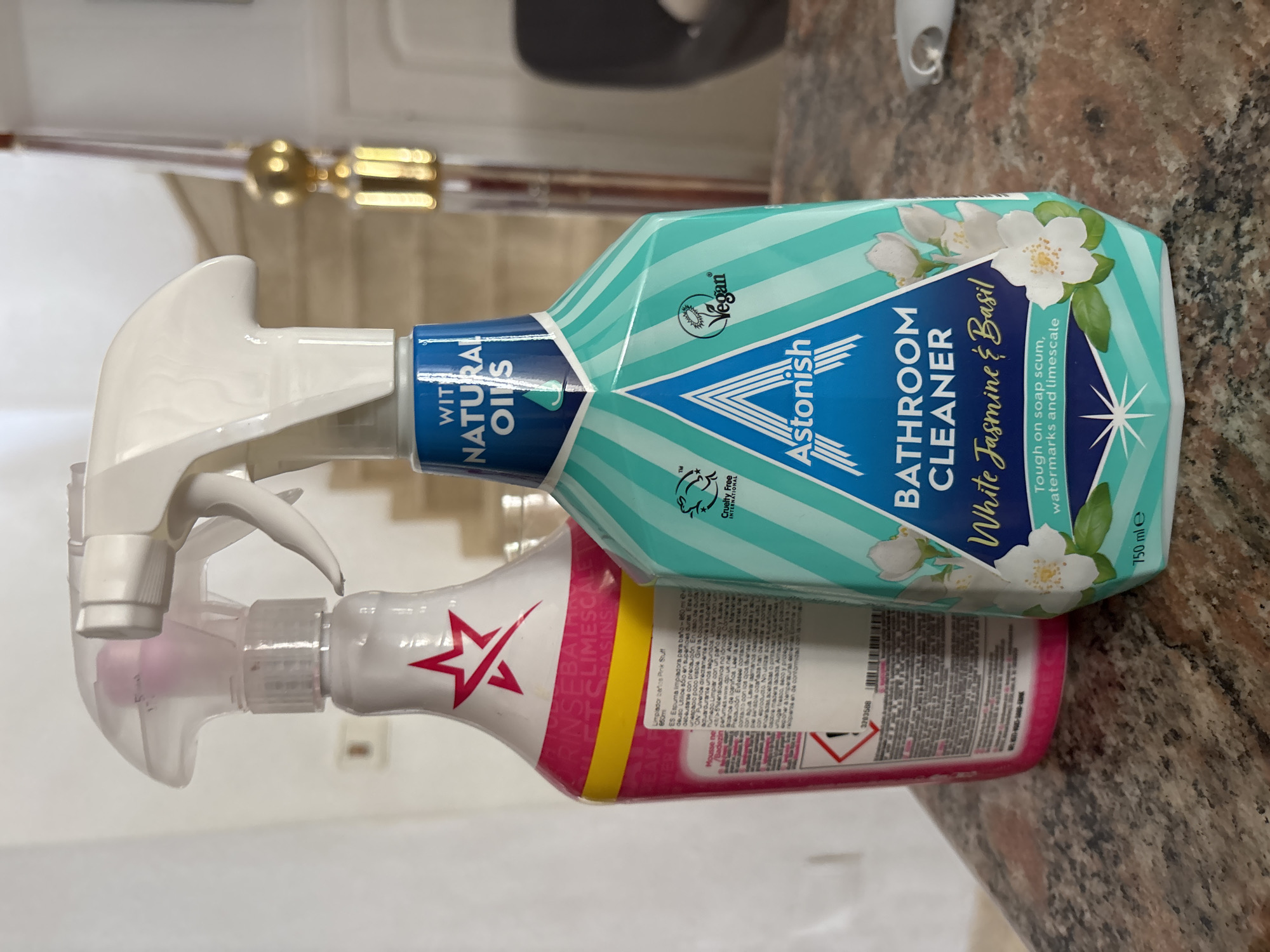 The image shows two bottles of cleaning product on a granite countertop in what appears to be a domestic setting, possibly in the city of Orihuela, Spain. The bottle on the right is an "Astonish Bathroom Cleaner" with a White Jasmine & Basil scent, featuring a teal and white striped design with floral imagery and vegan and cruelty-free certifications. The bottle on the left is partially obscured but appears to be a pink cleaning product with a star logo, possibly "Rinse & Shine." In the background, a staircase with a gold balustrade is visible, along with a doorway. The lighting suggests an indoor environment, possibly with natural light coming from a window.