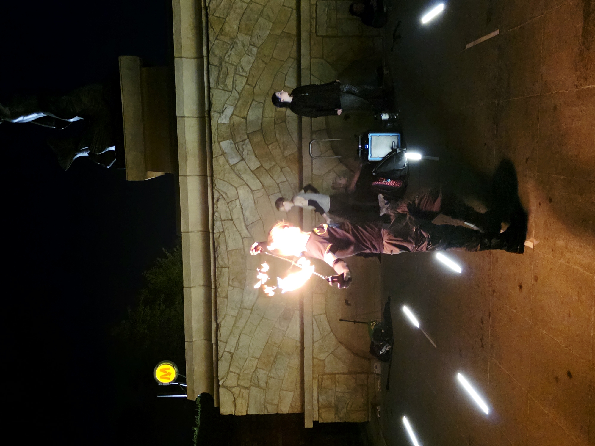 The image depicts a nighttime scene outdoors in front of a stone wall, with a statue visible above. A performer is in the foreground, twirling a poi prop that is on fire, illuminating their face and body with a bright orange glow. They are wearing dark clothing and black boots. Several people are standing and sitting in the background, observing the performance. To the left of the performer, a yellow and red circular sign with the letter "M" is mounted on a pole, likely indicating a metro station. The ground is paved with square tiles, and several illuminated white rods are placed on the ground, creating a light effect. Bags and a speaker are visible on the ground near the spectators. The overall ambiance suggests a public performance or event in Warsaw, Poland, given the presence of the metro sign.