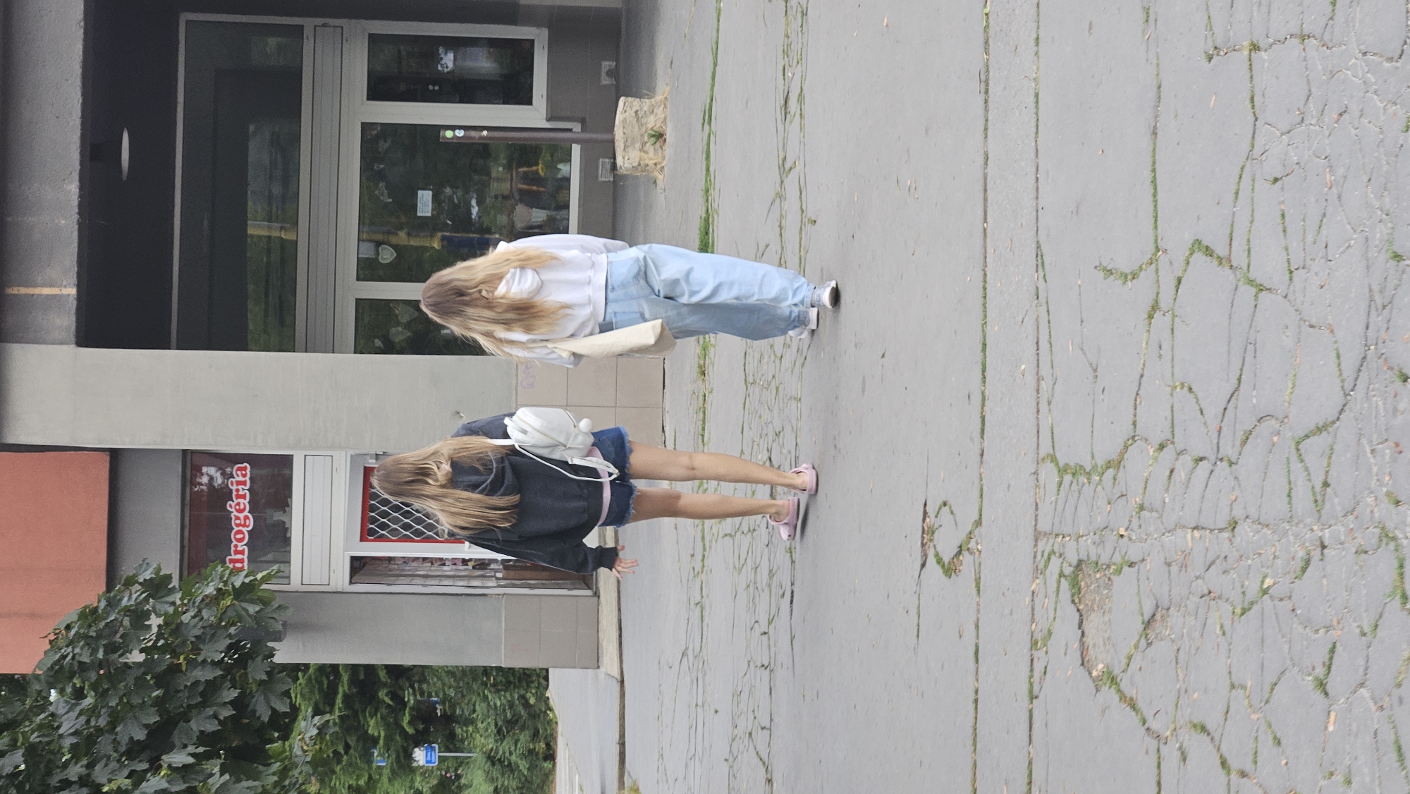 Two young women walk away from the camera on a cracked concrete sidewalk in front of a building. The woman on the left has long blonde hair and wears a dark hooded sweatshirt, denim shorts, and pink sandals. She carries a white backpack. The woman on the right has long blonde hair and wears a white hooded sweatshirt and light blue baggy jeans. She carries a light-colored tote bag. The building has a red sign that says "drogéria" in white letters. The scene appears to be in the city of Prešov, Slovakia.