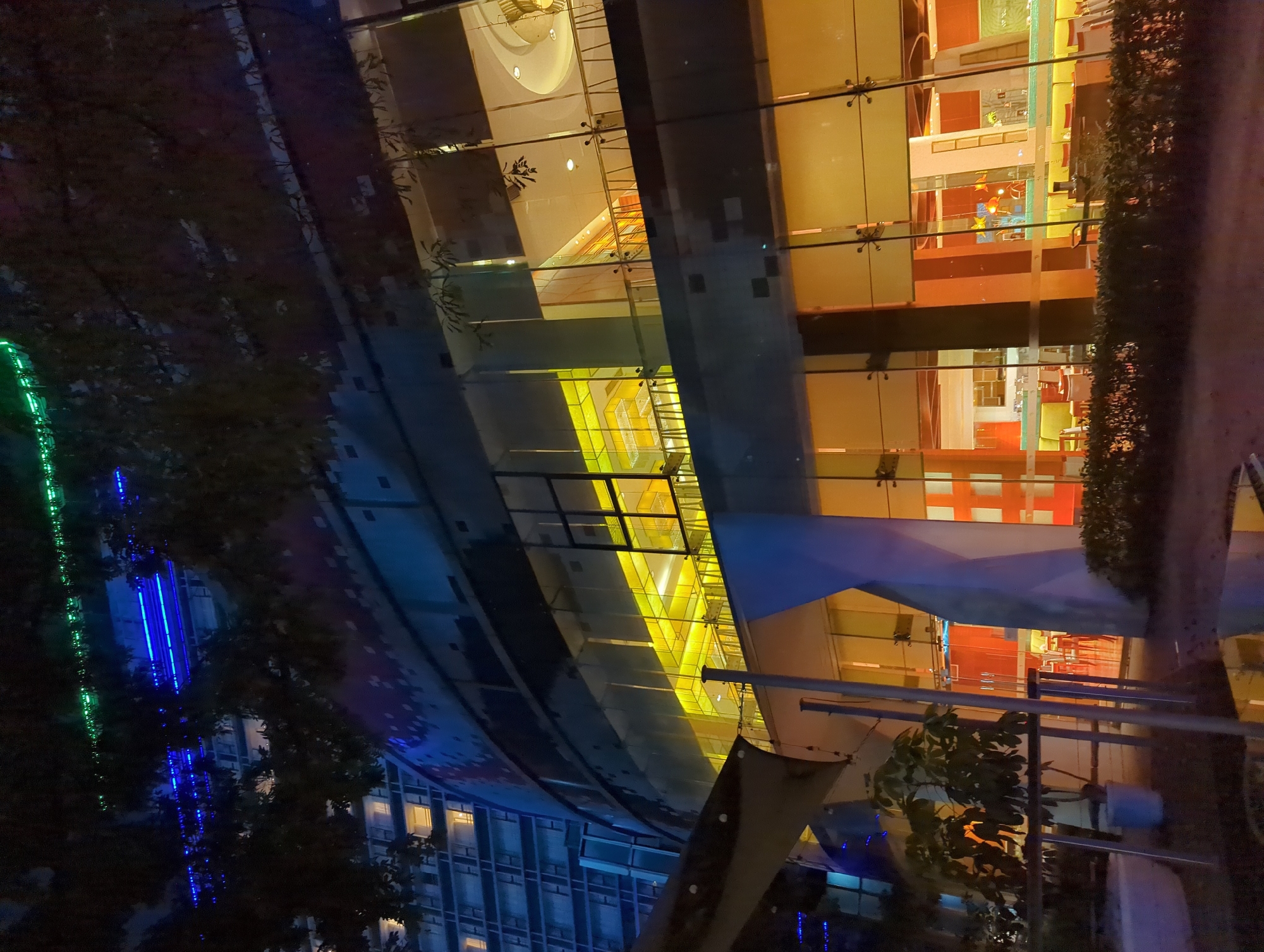 This is a nighttime, medium-shot, exterior photograph of a modern, multi-story building in Wuhou District, China. The building features a curved facade with extensive glass windows. Blue and green neon lights are visible along the top of an adjacent building, and tree branches with sparse foliage are in the foreground, obscuring parts of the view. The glass walls of the building reveal brightly lit interior spaces, including what appears to be a restaurant or lounge area on the upper floors with yellow and orange lighting. Below, a ground-level entrance area is visible, with red-toned accents and glass doors. The foreground shows a patch of dark ground with some reflective puddles, suggesting recent rain, and a low hedge. The scene is dark, with artificial lighting being the primary source of illumination.