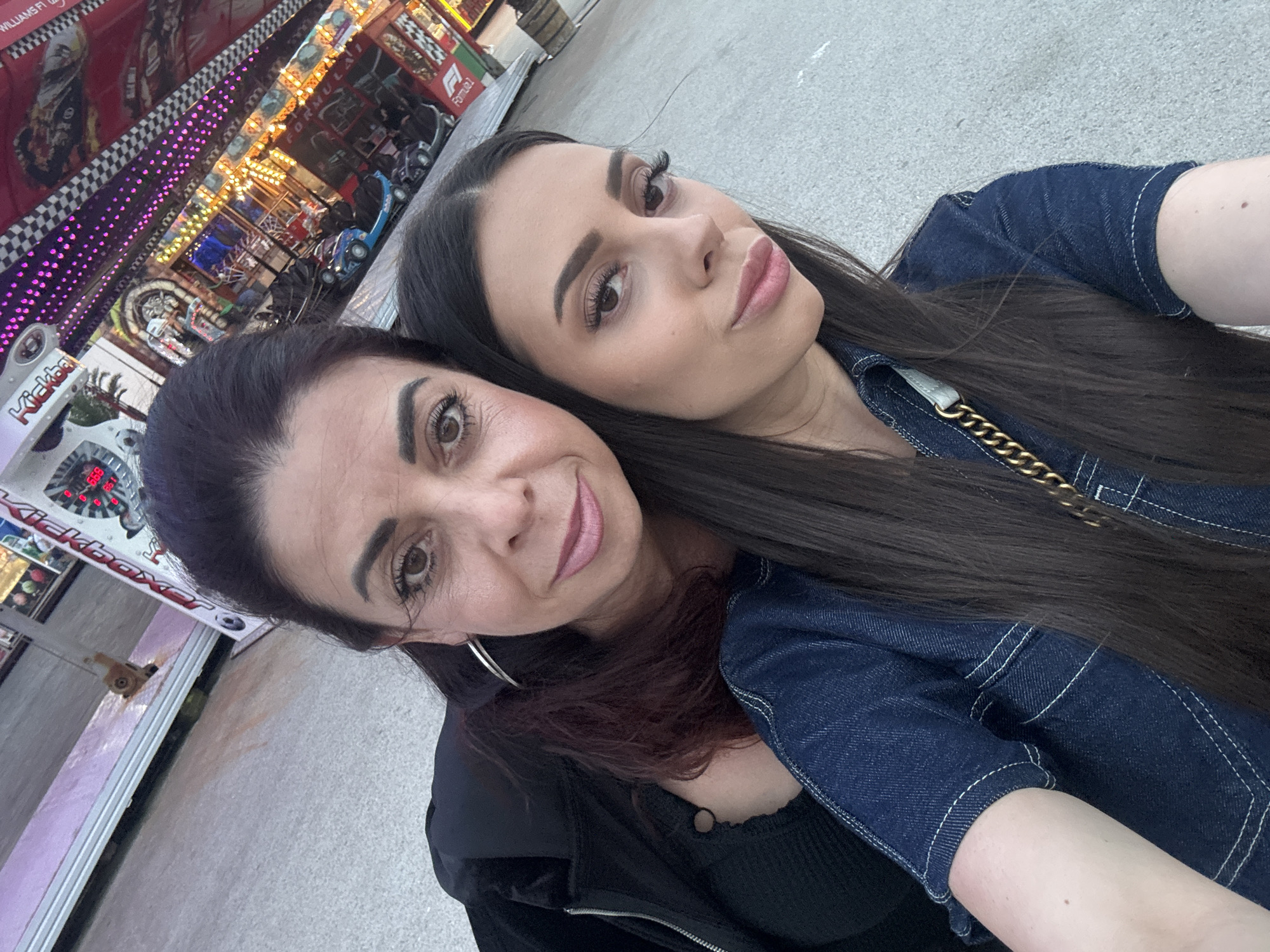 A selfie image, captured in Varna, Bulgaria, features two women positioned close together in the foreground. The woman on the left, appearing older, has dark hair with some auburn highlights visible on her shoulder, large silver-colored hoop earrings, and wears a black zippered jacket over a black ribbed top. She has defined eyebrows, dark eyelashes, and pink lipstick. The woman on the right, appearing younger, has long, straight dark hair parted slightly off-center and wears a dark blue denim short-sleeved top with white contrast stitching, with a gold-colored chain strap visible on her right shoulder. She also exhibits defined eyebrows, prominent dark eyelashes, and pink lipstick. Both women are looking directly at the camera. The background, angled and out of focus, reveals an amusement park environment. Visible elements include a "Kickboxer" ride sign with red digital displays on the far left, a "Formula 1" themed bumper car attraction with multiple bumper cars (some blue, some black), bright purple string lights, and yellow/orange illumination from the rides. A black and white checkered pattern is visible on an overhead structure, and a wooden barrel is partially visible on the far right. The ground beneath the individuals is light gray concrete.