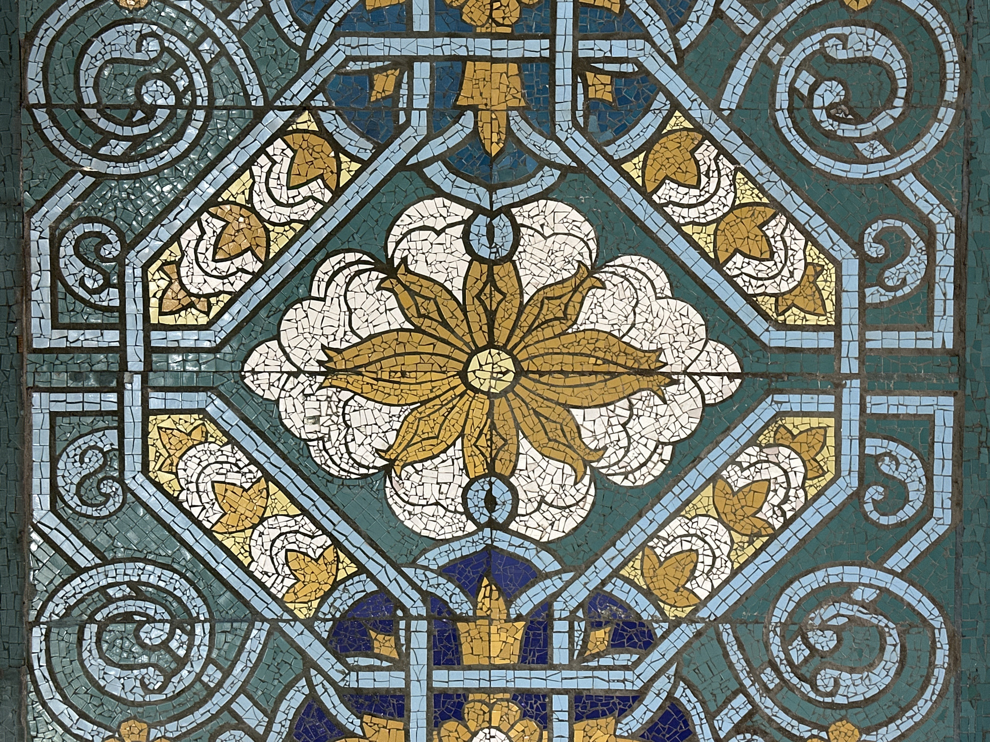 The image shows a close-up of a mosaic pattern. The mosaic features a central floral design with a golden center surrounded by white petals. The background is primarily composed of green, blue, gold, and white tesserae (small mosaic tiles), arranged in geometric and stylized floral shapes. The overall pattern suggests a repeating design element.
While the location isn't explicitly in the image, the prompt implies that the mosaic is in Tashkent, Uzbekistan. However, without additional context, it is not possible to pinpoint the specific location, or determine the time or whether the image is documentary in nature.