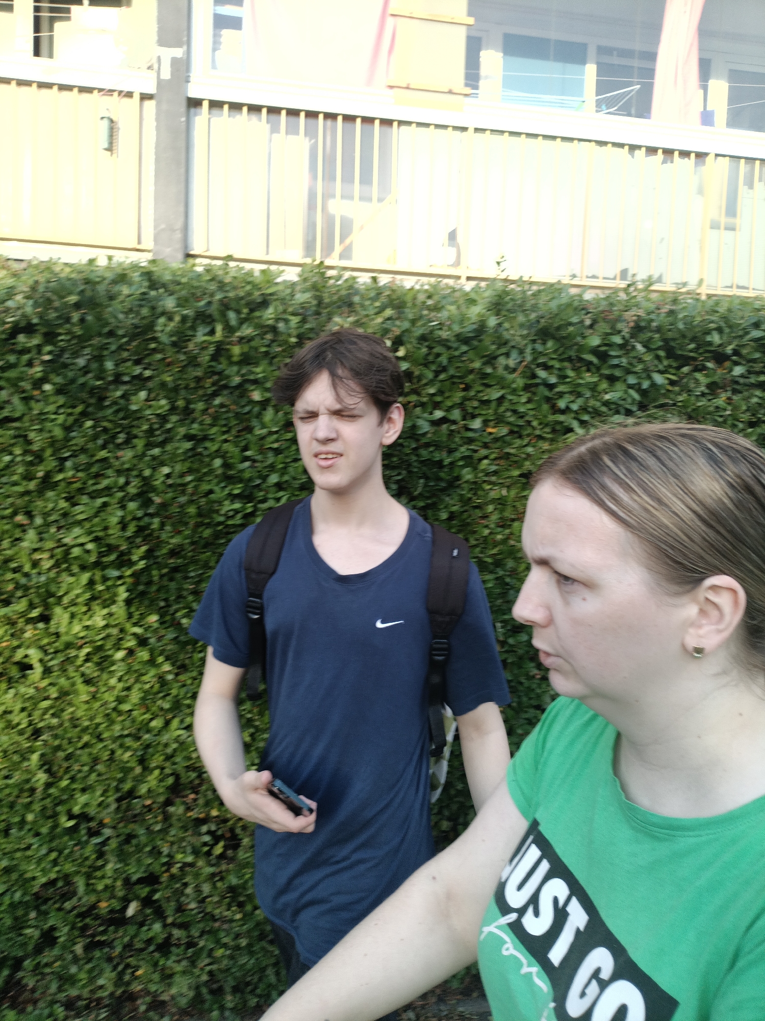 Two people are captured in a close-up, slightly blurred, outdoor medium shot. On the left is a young person with dark, messy hair, wearing a dark blue t-shirt with a white Nike logo. They are looking to their left with their eyes closed and mouth slightly open, and they are holding a dark-colored smartphone in their right hand. They are also wearing a black backpack. To the right is an adult with blonde hair, wearing a bright green t-shirt with the words "JUST GO" printed on it in white text within a black rectangle. They are looking towards the left, with their mouth slightly open. Both individuals are in front of a dense, dark green hedge. In the background, there is a building with light-colored vertical siding and horizontal grating. The lighting suggests it is daytime, with sunlight creating bright highlights on the building. No specific landmarks indicating Prague, Czechia are visible.