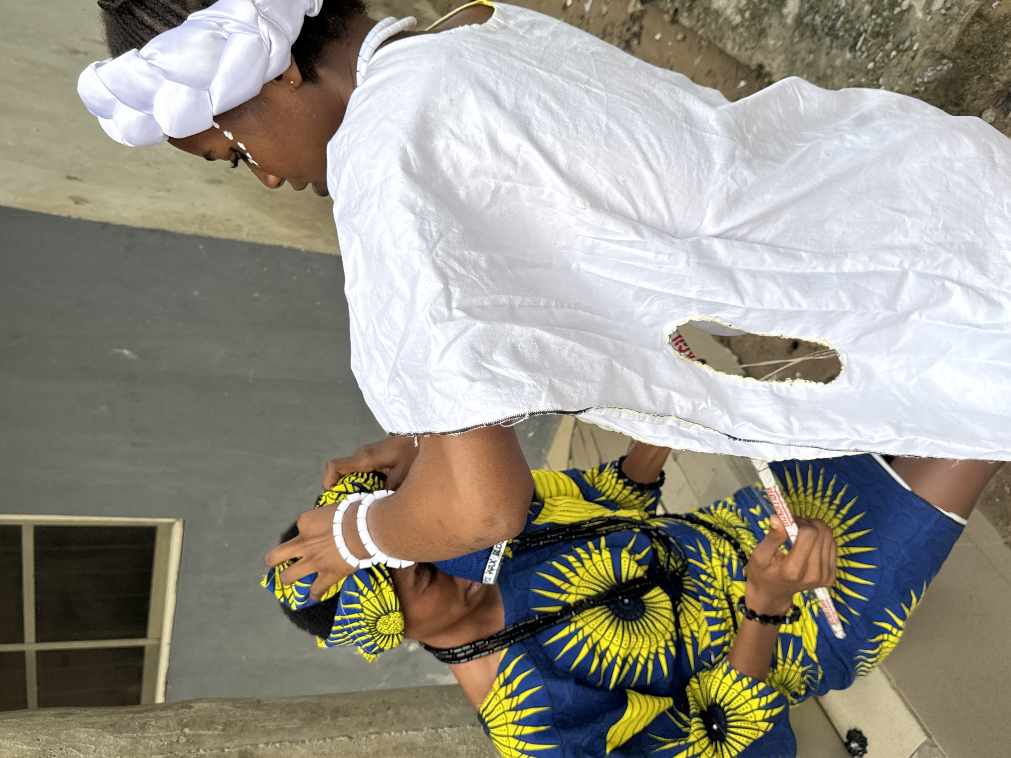 Two women are positioned near a concrete structure, likely outside. One woman wears a white outfit and head wrap, with what appears to be white paint on her face. The other woman wears a blue and yellow patterned outfit and head wrap, adorned with white bracelets and a black beaded necklace. She holds a white object in her hand. The environment shows a partial view of a building with a window. The location is speculated to be in Badagry, Nigeria.