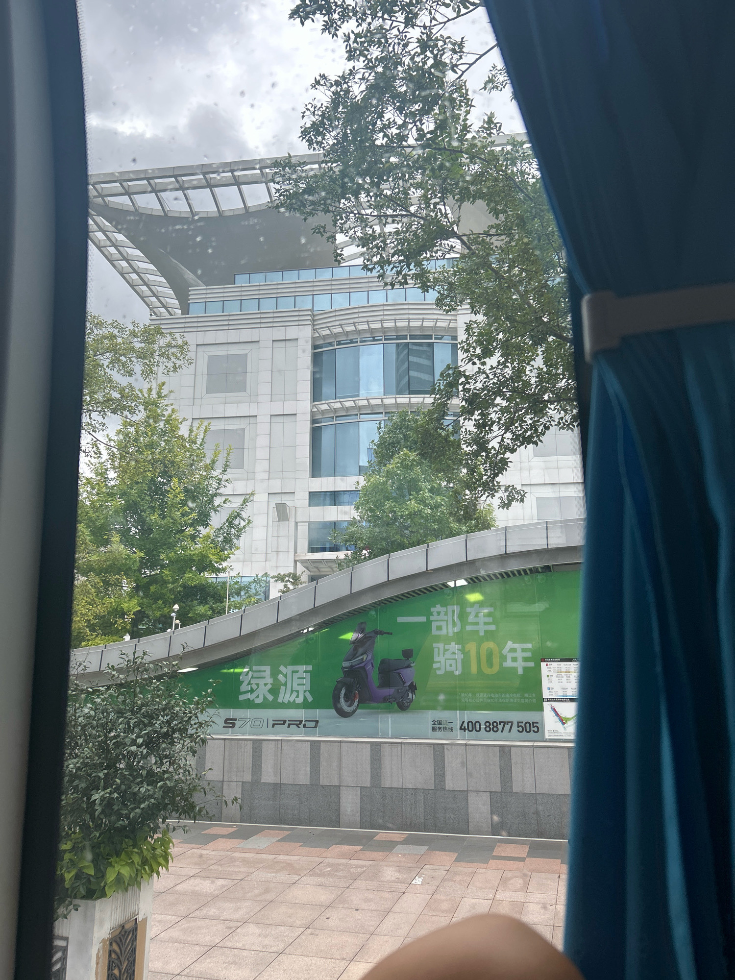 The image captures a view through a window, possibly from a vehicle, looking towards a modern building in Shanghai, China. A portion of a blue curtain is visible on the right side, framing the scene. In the background, a large building with a distinctive angular roof structure, composed of numerous geometric frames, dominates the upper portion of the frame. The building itself features multiple stories with large glass windows, reflecting the overcast sky.
In the middle ground, a large green billboard is prominently displayed, featuring an image of a purple electric scooter and Chinese text. The text on the billboard appears to be an advertisement. Below the billboard, a pedestrian walkway with patterned paving stones is visible. Several trees and some foliage are interspersed throughout the scene, adding a touch of greenery against the urban backdrop. The overall impression is that of a contemporary urban environment with commercial advertising integrated into the cityscape.
