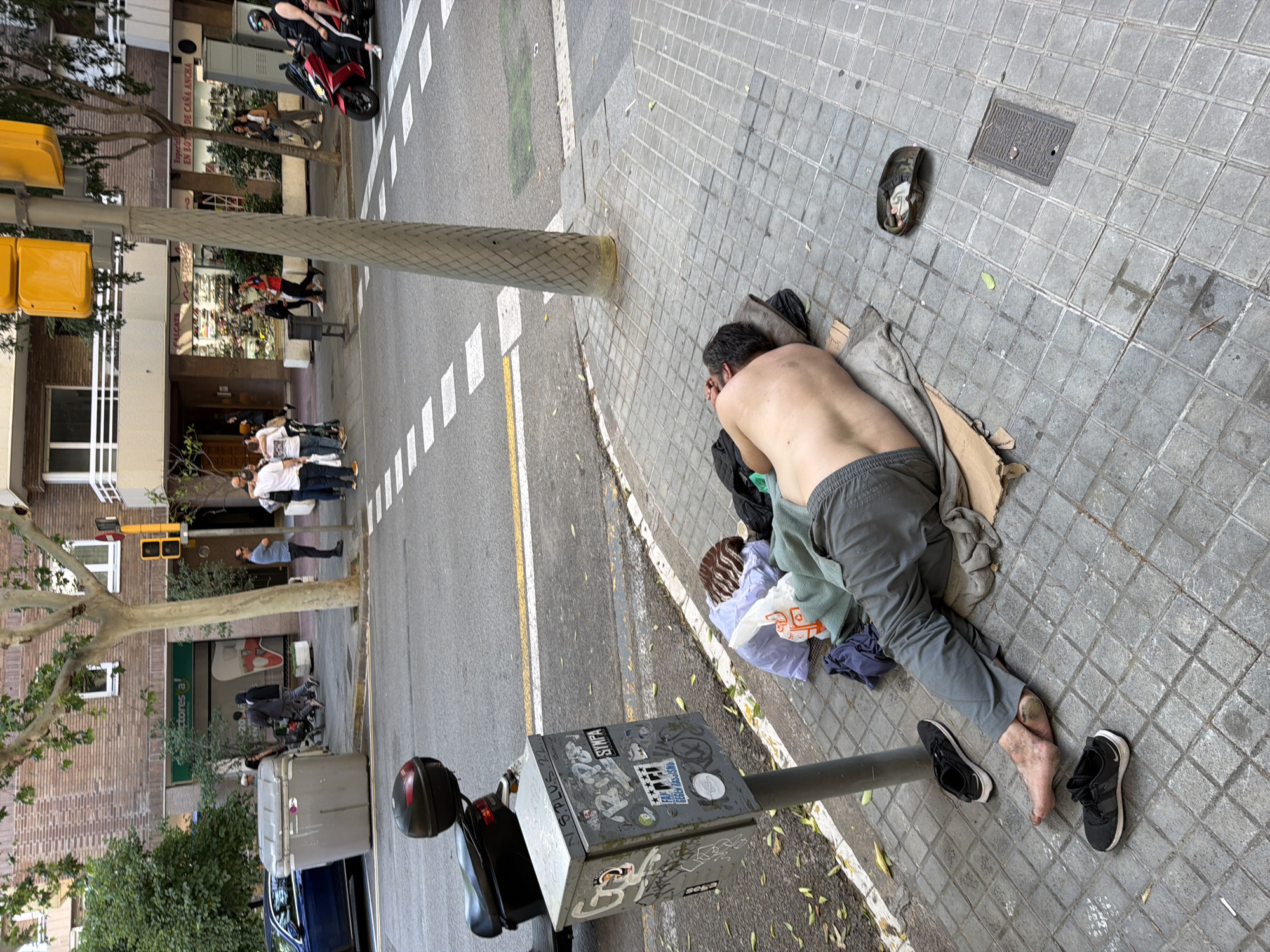 A shirtless man is lying on a sidewalk in Barcelona, Spain. He is wearing gray pants and black shoes, which are near his feet. A light-colored blanket and some clothing are near him. The sidewalk is paved with gray square tiles. A traffic light pole is next to him. A metal box with graffiti stickers is attached to the pole. The street is visible, with a crosswalk and a motorcycle. Buildings are visible in the background. A group of people is standing near a building with a green sign.
